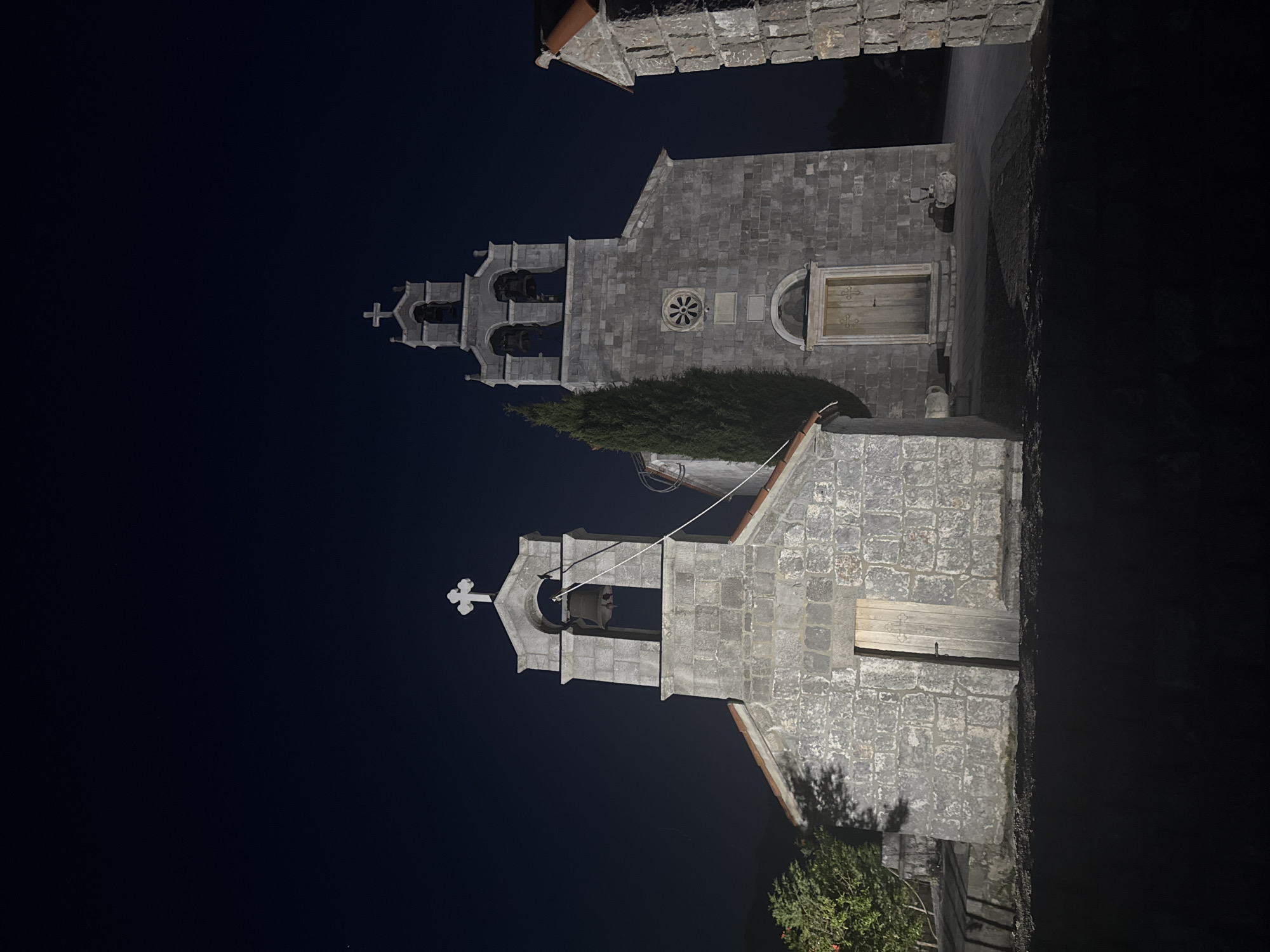 The image captures two stone church buildings in Kovačko Polje, Montenegro, at night. The sky is dark. The churches are illuminated. The building at the top of the image features a tower with three open archways, each containing a bell. Above the arches is a cross. Below the tower, there's a round window and a door set in an arched frame. A hedge partially obscures the view of the lower building, which is simpler in design, with a single bell tower topped with a cross. A rope is visible running from the upper part of this tower to the bell inside. Some vegetation is visible near the base of this building.