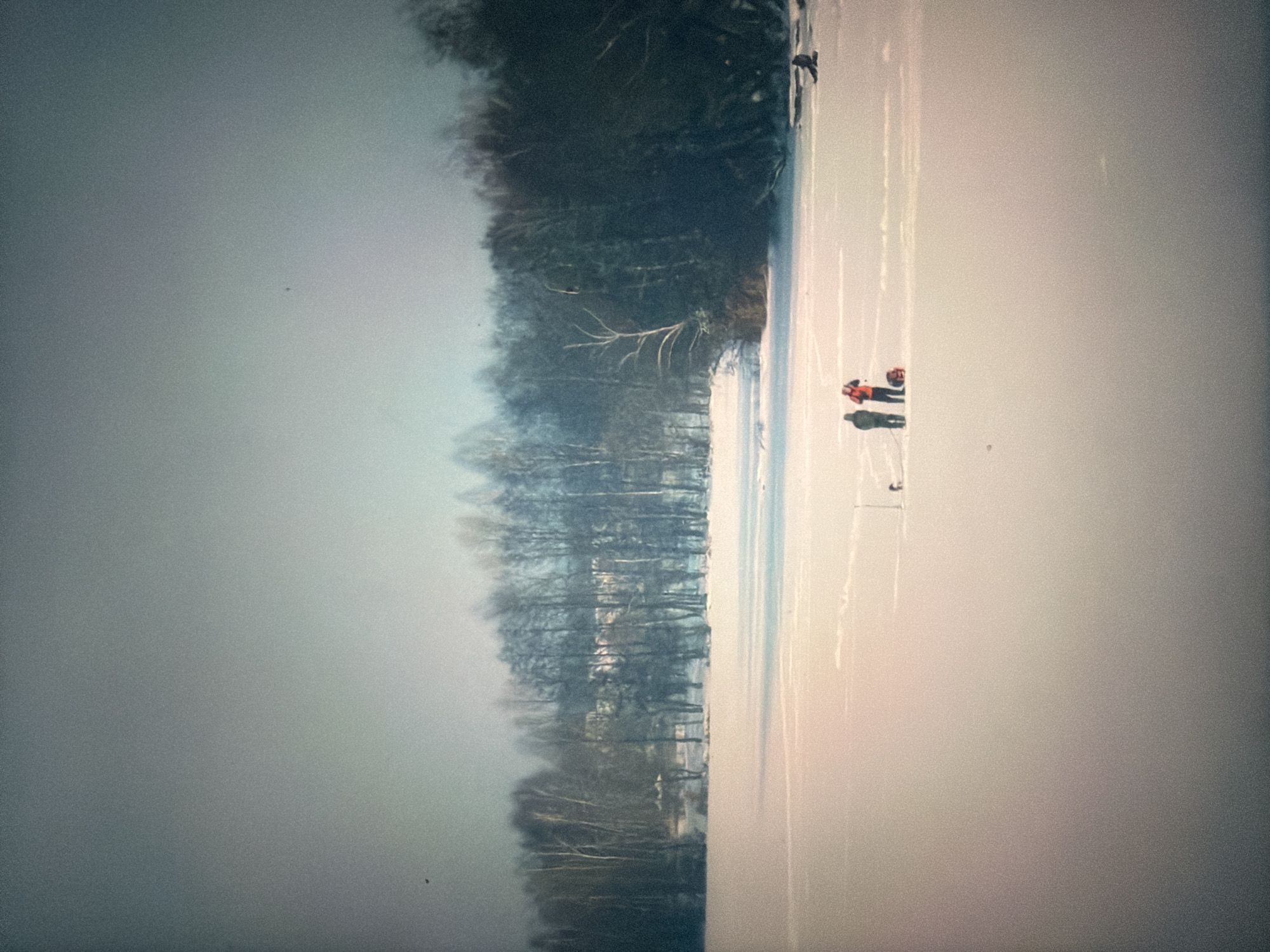 The image depicts a winter scene on a frozen body of water, likely a lake or river, with a treeline in the background. Two individuals are standing on the ice, one wearing a dark jacket and the other a bright orange jacket. A red bag or sled is positioned next to the person in the orange jacket. Further to the right, another individual is seated or lying down on the ice. In the distance, a forest of bare trees forms the horizon. The sky is overcast and a pale grey-blue. There is no identifiable information within the image to suggest a specific location in Krasnodar, Russia.