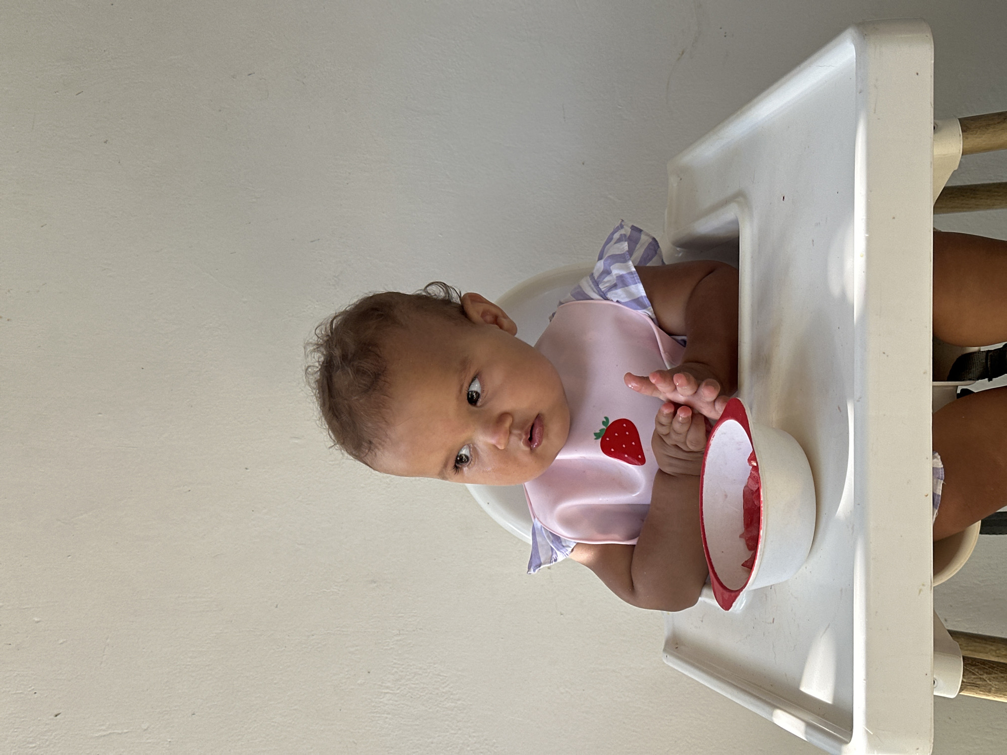 A young child sits in a white high chair in front of a plain, light-colored wall. The child, wearing a pink bib with a strawberry design and a purple and white striped shirt, looks to their left. In front of the child is a small white bowl with a red rim, containing several pieces of red fruit. The child's hands are clasped together on the tray of the high chair. The lighting suggests an outdoor or well-lit indoor environment, with shadows visible on the tray and wall. The location is identified as North Kuta, Indonesia.