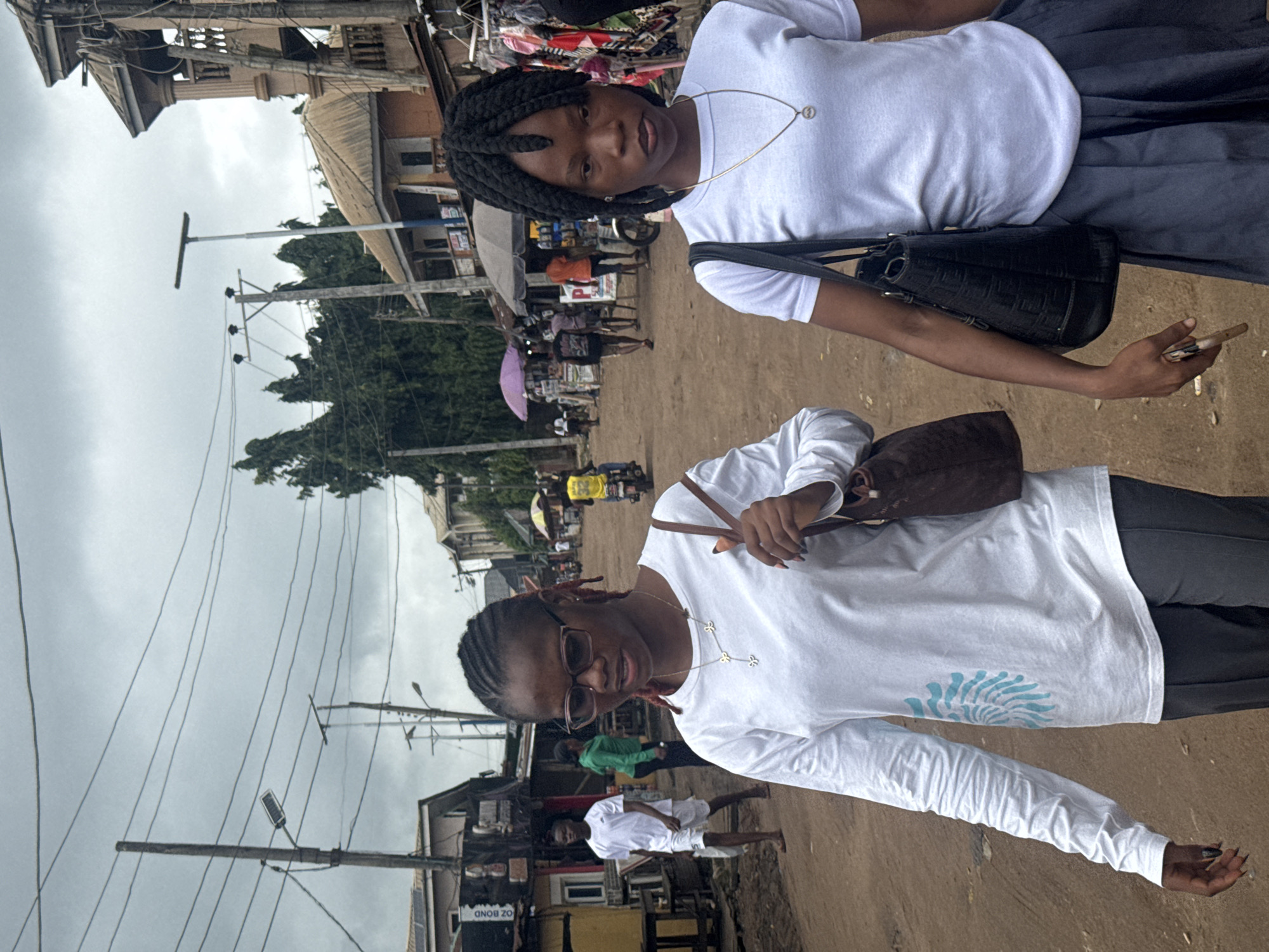 Two young women are walking on a dirt road in what appears to be a marketplace in Ihiagwa, Nigeria. The woman on the left is wearing a white long-sleeved shirt with a blue design and dark pants. She has glasses and is carrying a brown shoulder bag. The woman on the right is wearing a white short-sleeved t-shirt and a gray pleated skirt. She has dark braided hair and is carrying a black shoulder bag. In the background, there are several buildings, market stalls, and people. One man is walking in the distance wearing a white t-shirt and shorts. Another person is wearing a green shirt. Several motorcycles are visible, one of which has a rider wearing a yellow shirt. The sky is overcast.