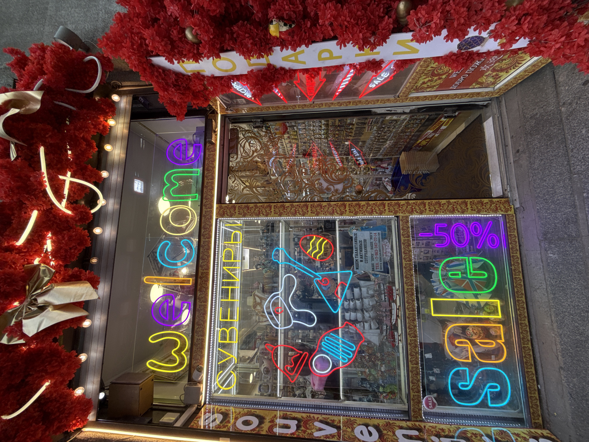This is a close-up, exterior shot of a storefront decorated with red artificial foliage and bright neon signs. The shop appears to be located in Saint Petersburg, Russia, as indicated by the Cyrillic text on one of the signs and the presence of a British flag motif in a neon sign.
The storefront features a large display window filled with merchandise, reflecting the neon lights and the surrounding decorations. The neon signs are a prominent feature, displaying text such as "Welcome," "Sale," "-50%," and Cyrillic words that likely translate to "Souvenirs" and "Welcome." Some of the signs also include abstract shapes and stylized graphics, including what looks like a guitar. The merchandise visible through the window includes various trinkets and items, possibly souvenirs, given the text on the sign.
The decorations suggest a festive or promotional event is taking place, with the red foliage and the "Sale" signs pointing to a possible holiday season or special offer. The lighting is vibrant and colorful due to the neon signs, creating a lively and eye-catching display against the more subdued background of the building's exterior. There are no people visible in the image. The context suggests the photo was taken during daylight hours, though the neon lights are still a primary source of illumination within the frame.