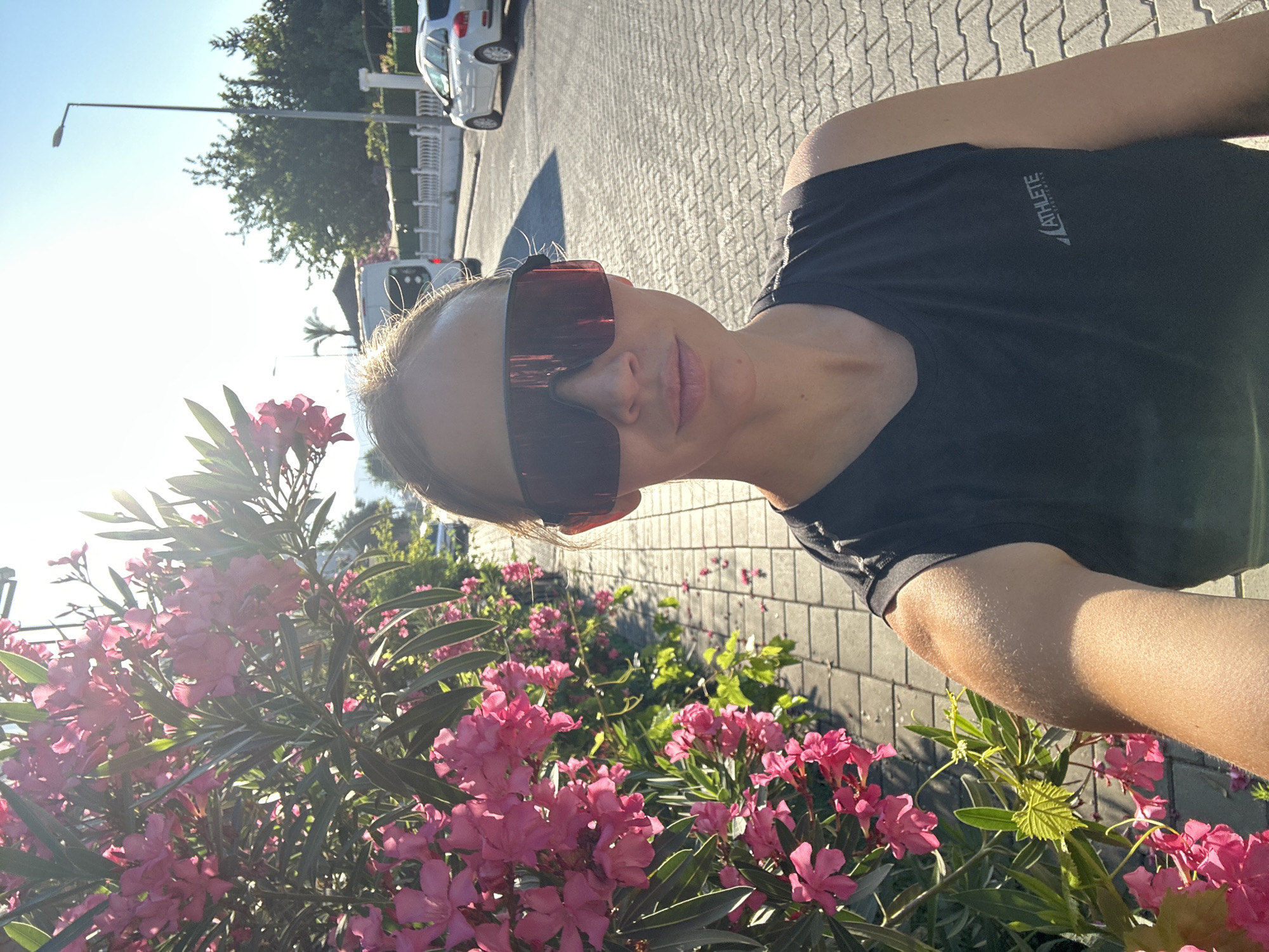 A woman is captured in a sunlit outdoor selfie, seen from the chest up, with her head tilted slightly. She wears a black t-shirt featuring the word "ATHLETE" and a stylized logo printed in white on the left side of the chest. Her light-colored hair is pulled back, and she is wearing large, dark, rectangular sunglasses that obscure her eyes. Her lips are slightly parted, and she has a neutral expression.
The foreground and left side of the image are dominated by a lush bush with numerous vibrant pink flowers and long, narrow green leaves, typical of Oleander. Bright sunlight streams through the foliage, creating a strong glare and indicating a clear, sunny day, likely in the morning or late afternoon due to the low angle of the sun and prominent shadows visible on the ground.
Behind the woman, a paved area with a distinct cobblestone-like pattern stretches out. In the background, parked cars, including a silver SUV and a white van, are visible along a street. Trees, including a distant palm tree, and a lamppost line the thoroughfare. The overall setting is an outdoor street or pathway in a warm, Mediterranean-like environment, consistent with Fethiye, Turkey.
