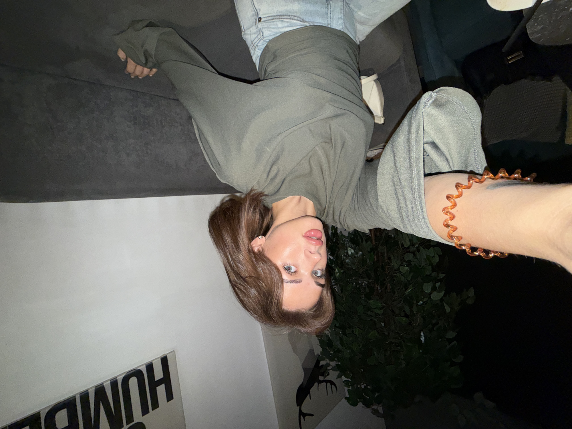 A woman with brown hair and light-colored eyes is lying upside down on a gray couch. She is wearing light blue jeans and a long-sleeved, olive-green shirt. Her left arm is outstretched towards the camera, displaying a spiral orange bracelet on her wrist. Behind her, a white wall is visible with black lettering and a black deer silhouette. A dark green leafy plant stands near the wall. The background appears to be dark, possibly indicating nighttime. No specific location is determinable from the image.