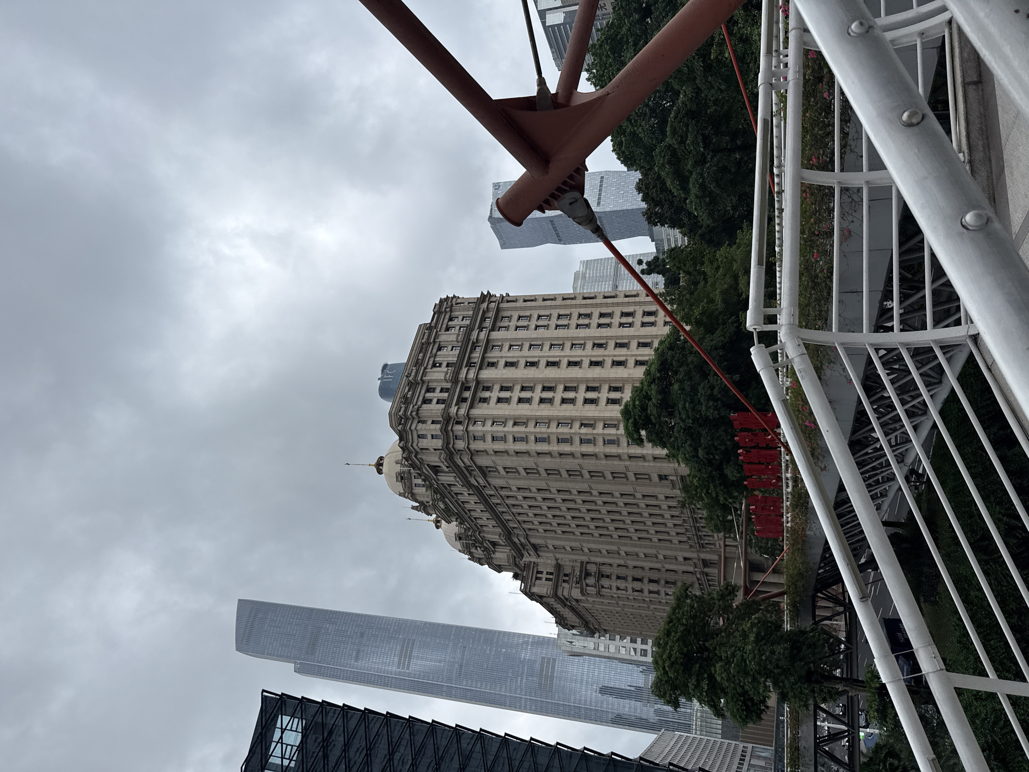 A low-angle, wide shot captures a cityscape in Tianhe District, China, dominated by a mix of modern skyscrapers and a historic building. The sky is overcast and gray. In the foreground, a section of a white pedestrian bridge with metal railings is visible, leading the eye towards the city. Behind the bridge, a grand, ornate, beige building with multiple stories and domed roofs stands prominently. To its left, a sleek, very tall, glass skyscraper reflects the cloudy sky. To the right and further back, another modern skyscraper with a unique twisted design is visible. Lush green trees partially obscure the lower portions of the buildings and bridge structures. Red banners or decorations are strung across an elevated walkway in the middle ground, adding a splash of color. The overall scene depicts an urban environment with a blend of architectural styles.