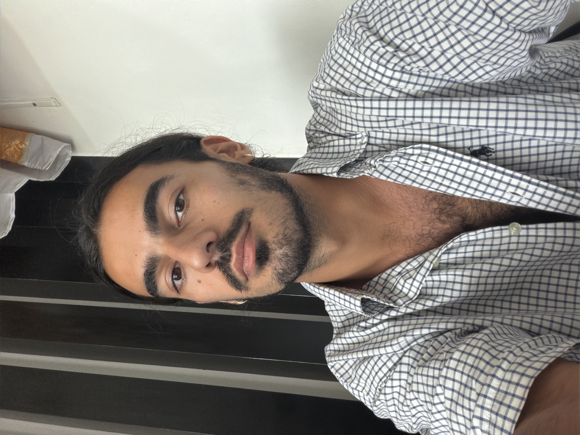 A man with dark hair and a beard is wearing a white and blue checkered shirt. The shirt is partially unbuttoned, revealing some chest hair. He has an earring in his left ear. The background consists of a white wall and a black surface with horizontal lines. There are some personal effects in the upper left, out of focus. This image has no features to identify Mengwi, Indonesia.