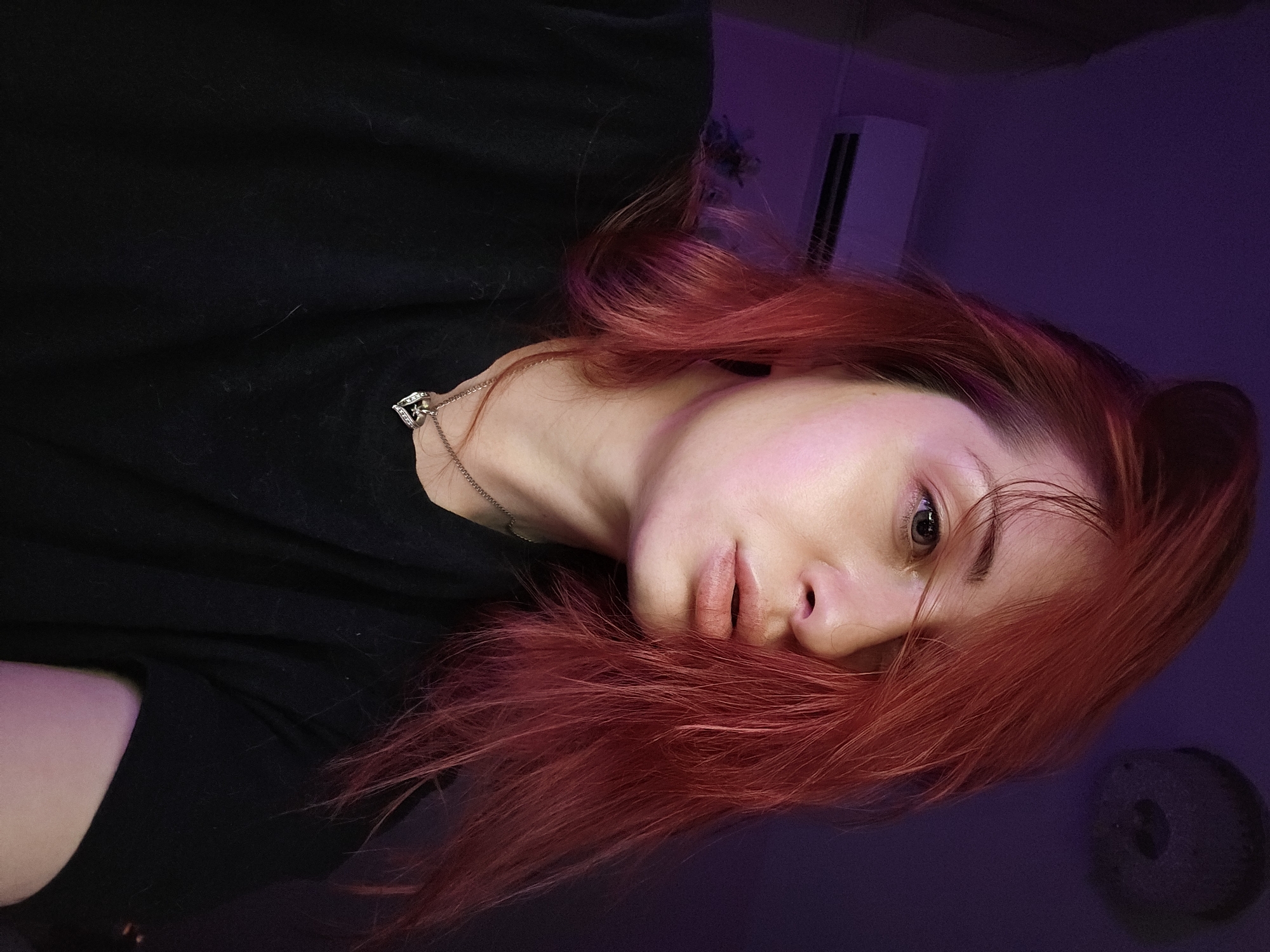 A female individual with long, reddish-auburn hair, wearing a black short-sleeved top and a silver chain necklace with a heart-shaped pendant, is depicted in a close-up, chest-up shot. Her face, characterized by fair skin, is illuminated by a prominent purple-pink light source, casting a distinct hue. Her left eye is partially obscured by hair, while her light-colored right eye is fully visible and directed slightly off-camera to her right. Her lips are slightly parted. The background is a dark purple wall. An air conditioning unit is mounted on the wall in the upper left portion of the image, and a dark, circular ceiling light fixture is visible in the upper right. The overall lighting is dim and predominantly artificial. There are no discernible visual elements or landmarks within the image that identify the specific location as Burgas, Bulgaria.