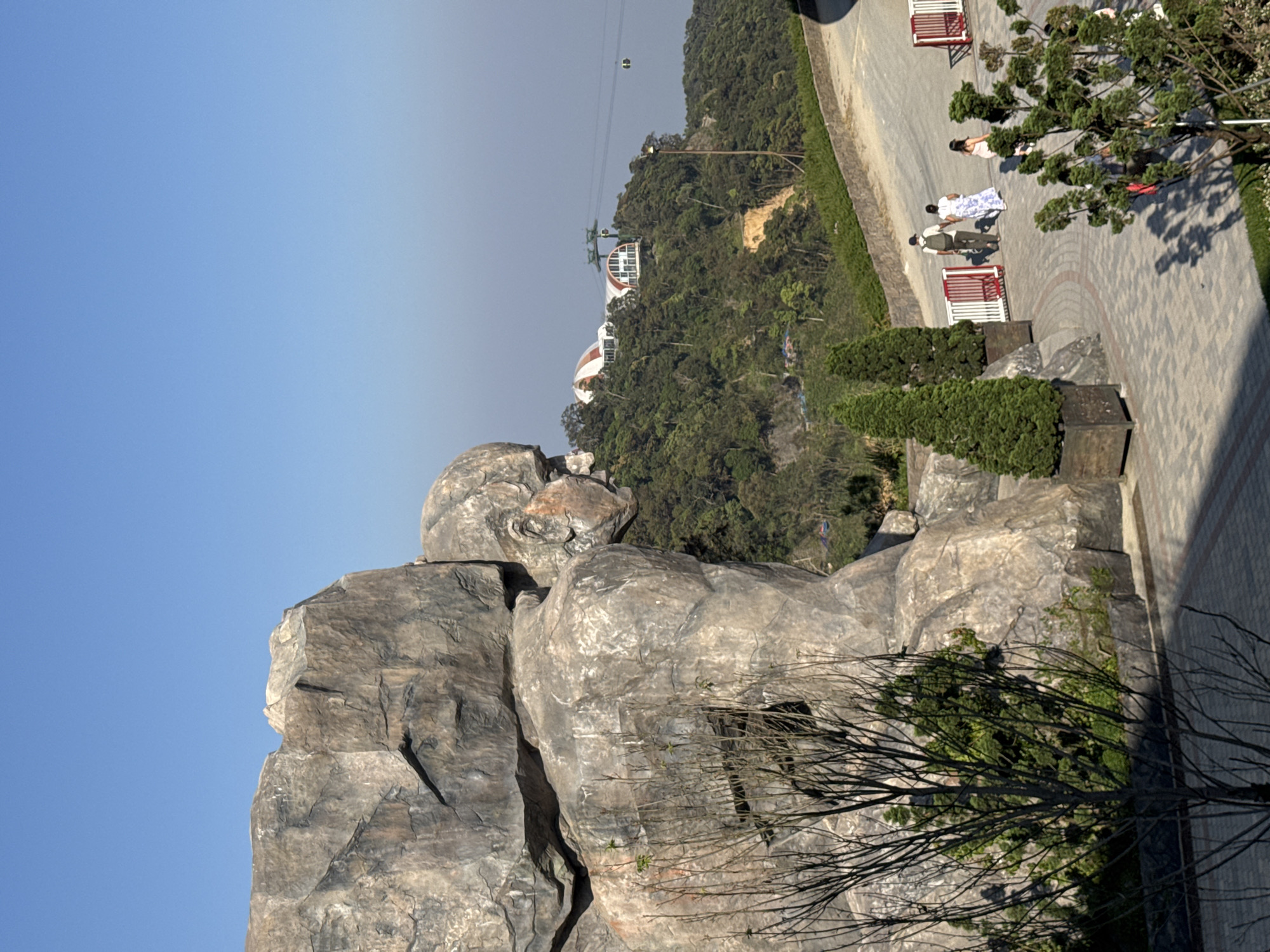 This image depicts an outdoor scene at Ba Na Hills SunWorld, located in Đà Nẵng, Vietnam. The primary subject is a massive, sculpted, grey-toned rock formation resembling a crouching or seated human figure in profile, facing right, positioned prominently on the left side of the frame. Its surface exhibits artificial rock textures, including cracks and varied shading. Bare branches of a deciduous tree partially obscure the lower section of the rock formation in the immediate foreground.
In the middle ground, a wide, curving pathway of light grey and reddish-brown interlocking pavers extends from the left to the right. Four individuals are observed walking on this pathway from left to right: a male wearing a light-colored hat and backpack walking beside a female in a long white and blue patterned dress, followed by a second female in a light pink dress, and a third female partially obscured by foliage further right. Two tall, conical evergreen trees in dark brown planters are positioned along the left edge of the pathway, and multiple white and red barriers line its perimeter.
The background consists of densely forested hills. Two buildings with distinct white and red-arched roofs are visible on a distant hillside. A cable car system with green support pylons and at least two moving gondola cabins traverses the sky above the hills. The upper portion of the frame is a clear, bright blue sky, indicating sunny weather. Strong natural sunlight casts distinct shadows from the trees and individuals onto the paved pathway.