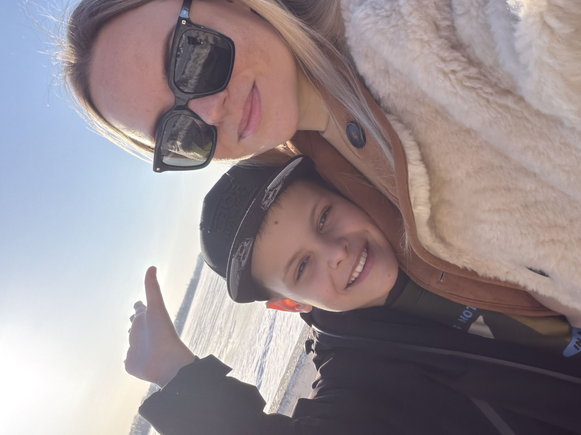 Two individuals are pictured outdoors in Votkinsk, Russia. One appears to be a woman with blonde hair and sunglasses, wearing a light-colored fur jacket over a brown garment. The other is a younger person wearing a dark cap and jacket. They appear to be taking a selfie. The background shows a frozen, snow-covered landscape under a bright sky, indicating it could be a winter scene. A raised thumb is visible in the foreground.