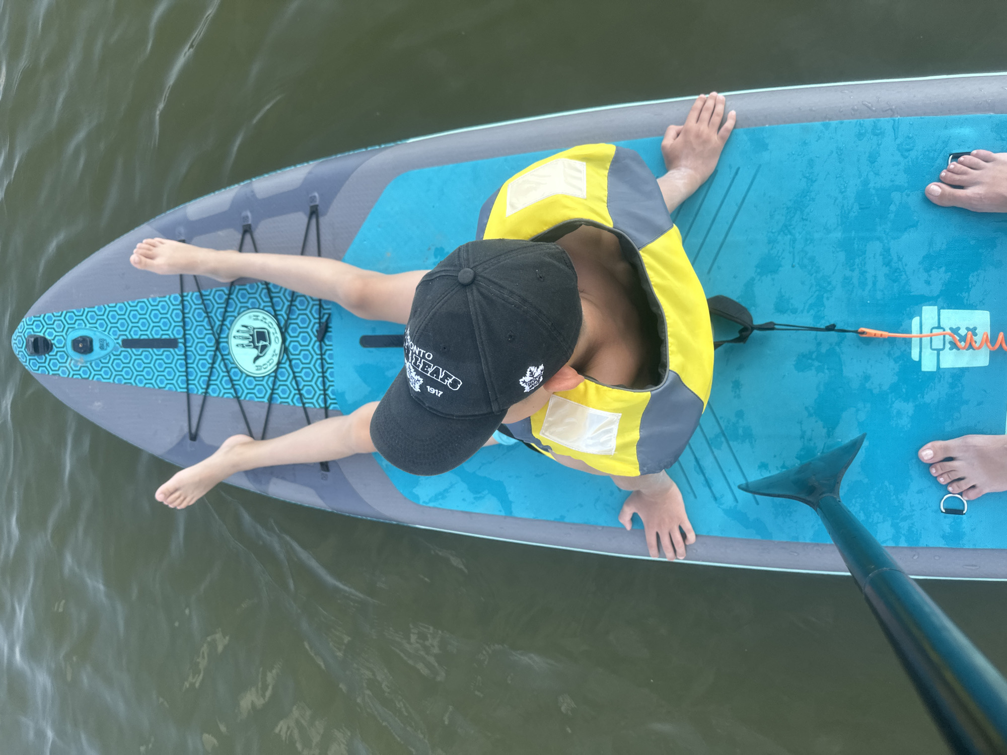 An overhead shot shows a person on a paddleboard in calm, murky green water. The person is wearing a black baseball cap, a yellow and grey life vest, and dark shorts. Their legs are extended forward, and their feet are visible at the front of the board. Their hands are resting on the sides of the paddleboard. A paddle is visible at the bottom left of the frame, with its blade angled towards the person. The paddleboard is blue and grey with a honeycomb pattern and a Body Glove logo. The water appears still with slight ripples.