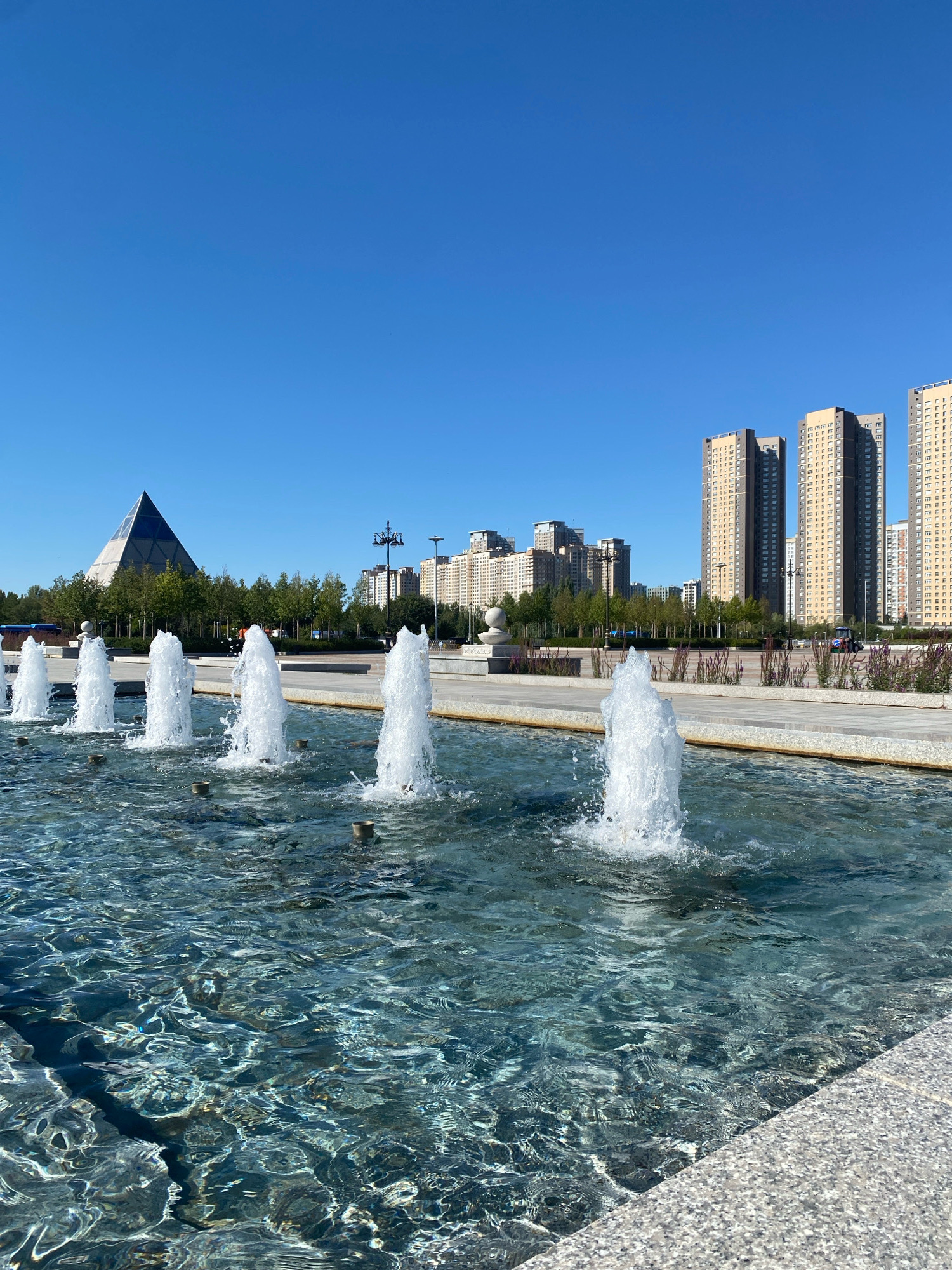 In Saraişyq District, Kazakhstan, a clear blue sky presides over an outdoor scene featuring a row of active fountains in the foreground. The fountains spray white water plumes into a large, shallow pool with rippling, clear blue water. The pool's edge is lined with a textured gray material. Beyond the fountains, a paved walkway is visible, bordered by sparse vegetation and decorative grass. In the background, a distinctive pyramid-shaped building stands to the left, its glass surface reflecting the sky. Further back, a line of trees separates the foreground from a cluster of tall, modern apartment buildings with numerous windows. The overall environment suggests a public space or park within a city.