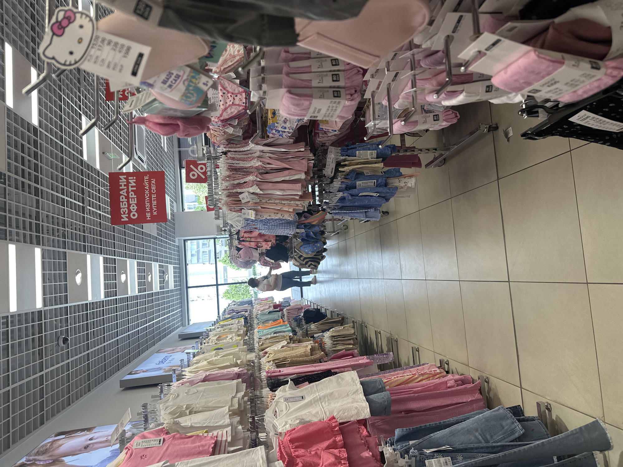 The image depicts the interior of a retail clothing store, likely a children's clothing section, situated in Panagyurishte, Bulgaria. The scene is characterized by rows of clothing racks filled with apparel for infants and children, predominantly in pastel colors like pink, white, and light blue. The merchandise includes dresses, shorts, shirts, and what appear to be undergarments or swimwear. Several price tags and promotional signs are visible, including one in Cyrillic that reads "ИЗБРАНИ ОФЕРТИ!" (Selected Offers!) and another with a percentage symbol. A woman, wearing a light-colored top and dark trousers, is browsing the racks in the background. The store environment features a suspended grid ceiling with fluorescent lighting and large windows at the far end, offering a glimpse of the exterior with trees and buildings. The flooring is tiled with a light beige color.
