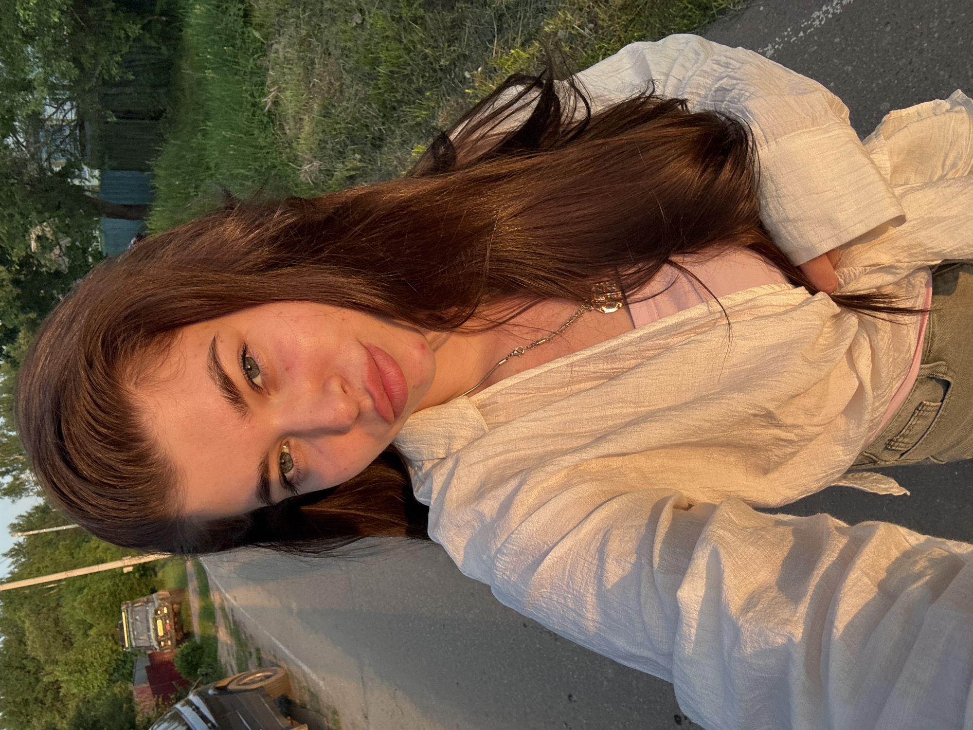 A young woman with long brown hair and light eyes is taking a selfie outdoors. She is wearing a white shirt over a pink top. A thin silver necklace is visible around her neck. The background shows a paved road, grass, trees, and buildings. The setting appears to be a residential area. Given the prompt, this location is assumed to be somewhere in the city of Мещёры, Russia. The lighting suggests it is late afternoon or early evening.