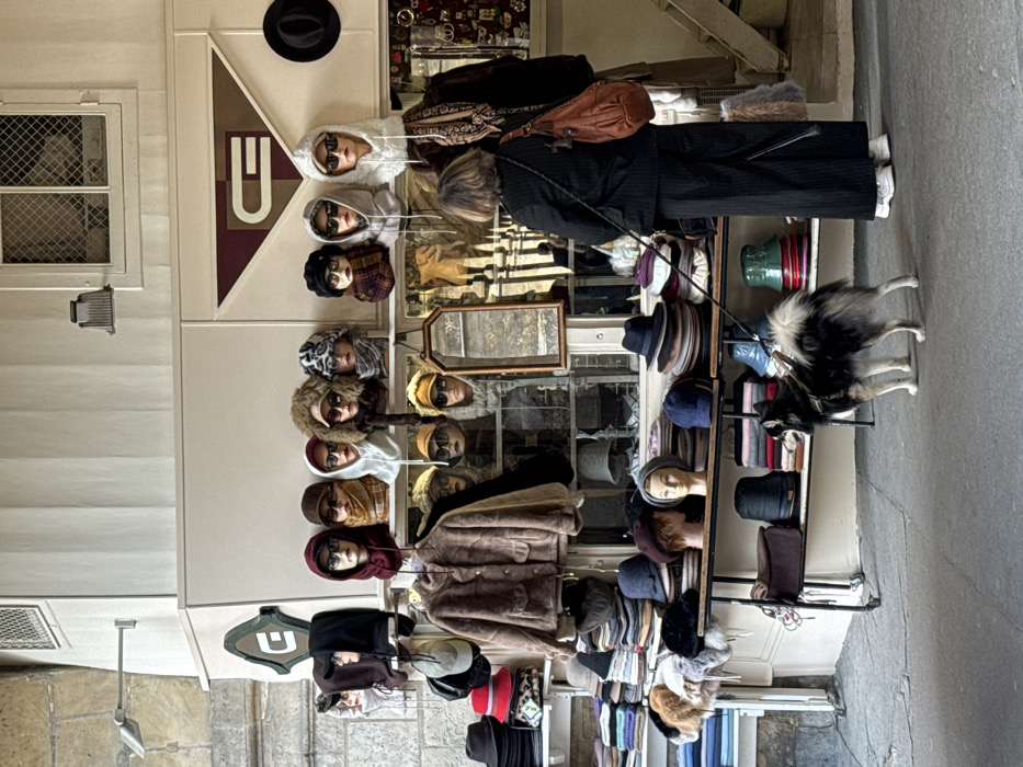 The image captures a detailed view of a hat and accessory shop situated within a covered pedestrian passage in Paris, France. The shop's cream-colored facade, featuring white trim, is visible on the right, with a window containing a security mesh and two wall-mounted light fixtures above the main display. Stylized "C" or "G" logos in green and brown adorn the facade. To the left, an old stone block wall with an arched architectural element defines the passage.
The shop's display consists of multiple tiers of merchandise. The uppermost level showcases at least twelve mannequin heads, each wearing a distinct hat, hood, or scarf, with several also sporting sunglasses or eyeglasses. A brown faux-fur jacket hangs on a central hanger on the mid-level, flanked by additional mannequin heads. A small, rectangular mirror with a wooden frame is positioned centrally in this section. Lower shelves and display tables are densely arranged with various hats, including fedoras, bucket hats, and knitted caps, alongside piles of folded textiles. A black fedora hangs on the far right wall, and small accessories are visible behind glass on the right side of the display.
On the grey paved floor in the foreground, a person is seen from behind, facing the shop display. They have medium-length, dark-blonde or greyish-brown hair and are wearing a dark, possibly pinstriped, jacket and wide-leg trousers, paired with white sneakers. A reddish-brown leather bag with a braided strap is slung over their right shoulder. The person holds a black leash in their left hand, which extends to a medium-sized Spitz-type dog standing to their right. The dog has a thick, black and white/cream coat, wears a black harness, and is looking towards the right.