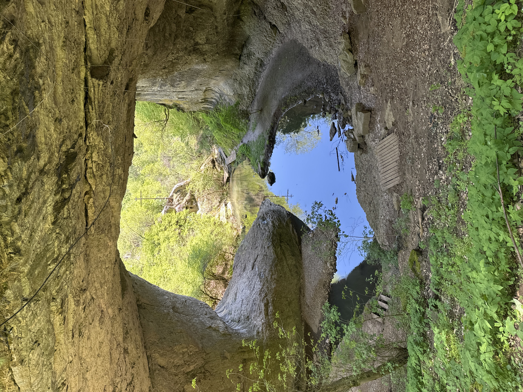 The image shows a view from inside a cave-like opening in Lilyache, Bulgaria, looking outwards. The walls of the opening are rough, light brown rock. Outside, there is a pool of water reflecting the sky, with surrounding green vegetation, trees, and rock formations. There is a small wooden platform next to the pool.
