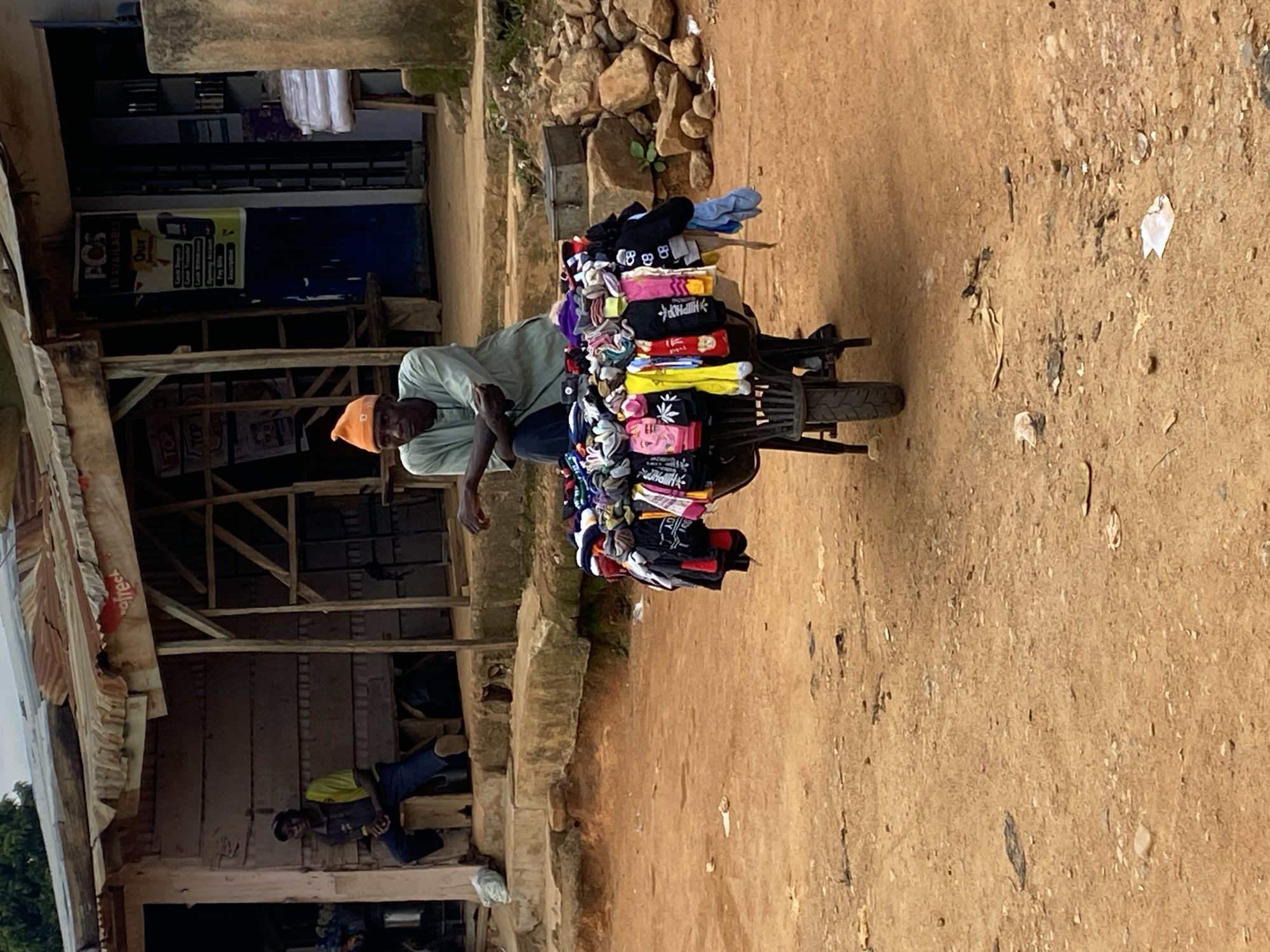 In the city of Gwong, Nigeria, a man wearing an orange beanie and a light green shirt sits on a stool behind a wheelbarrow filled with colorful socks. The wheelbarrow, a common mode of transport for vendors, is positioned on a dirt road. Behind the man, a structure resembling a shop or market stall is visible, with a corrugated metal roof and wooden supports. A second man, dressed in a yellow and dark blue shirt, sits on a step to the left of the stall. The environment appears to be a typical street scene in a Nigerian town, with a dusty road, rocks, and some vegetation. The overall impression is one of commerce and daily life in this West African setting.