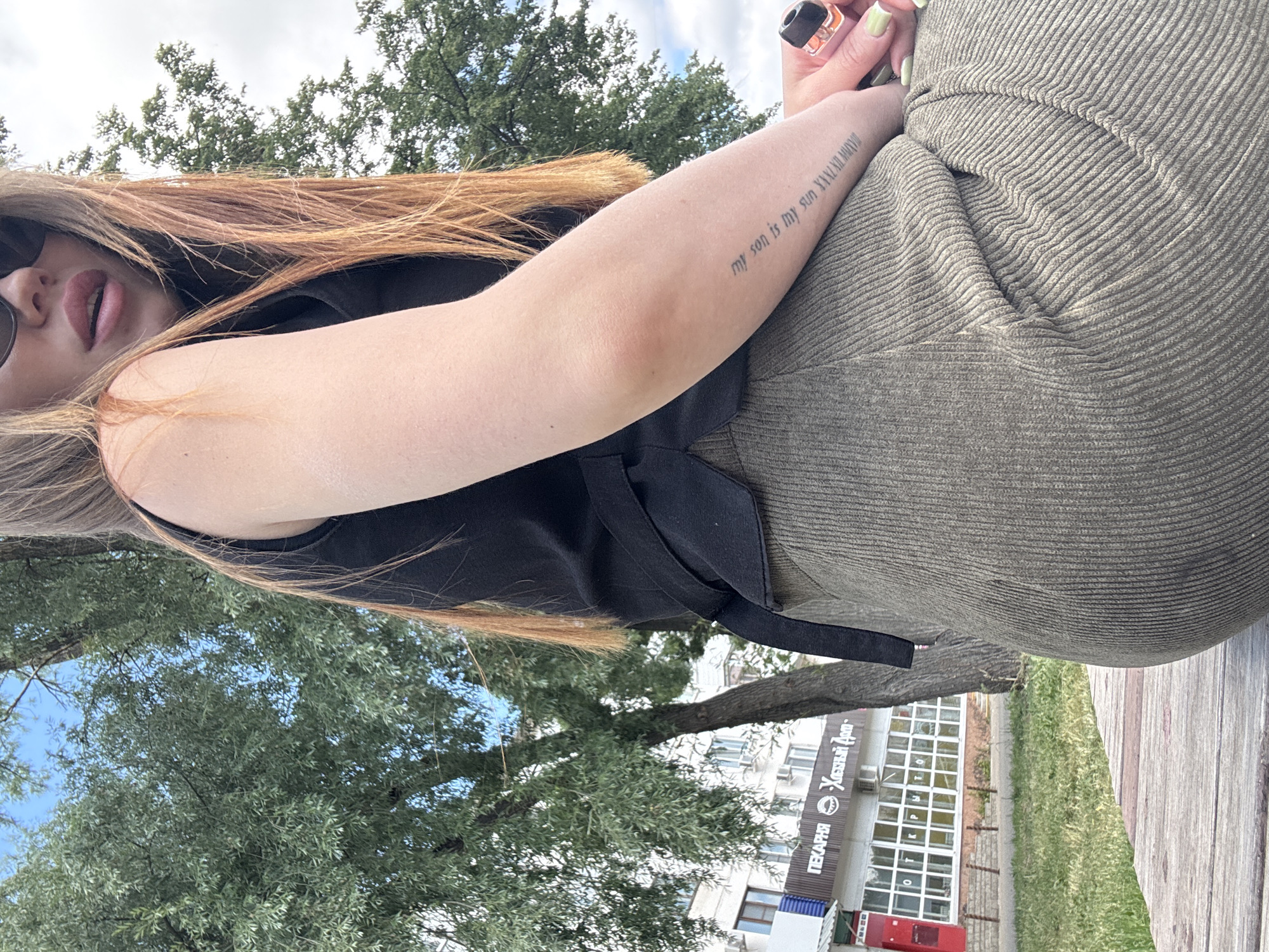 The image shows a young woman sitting on a wooden bench outdoors. She is wearing a black sleeveless top and a green ribbed skirt. A tattoo is visible on her left forearm, which reads "my son is my sun". She is also holding a vape device in her right hand. In the background, there is a tree, a building with signs in Russian, and a cloudy sky. The building has the text "ПЕКАРНЯ" and "Хлебный Дом" on it, indicating a bakery. The location is the city of Chaikovsky, Russia.