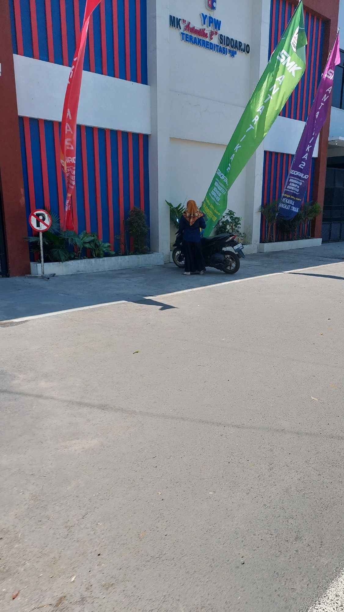 This image captures a sunny day outside a school building in Siwalan Panji, Indonesia. The building features a facade with alternating red and blue vertical stripes, and a white horizontal band. Above the entrance, text reads "YPW MK ANNAULHA SIDOARJO TERAKREDITASI". A tall, red banner hangs on the left side of the building.
In front of the building, a woman wearing a hijab and a dark outfit stands next to a black motorcycle. She is facing away from the camera, looking towards the building. Two large, colorful banners are prominently displayed: a vibrant green one on the left and a purple one on the right. Both banners have text that appears to be related to the school and its programs. The green banner has the text "SMK" and "SIDOARJO" visible, along with other Indonesian words. The purple banner also shows "SMK" and "SIDOARJO", and seems to highlight attributes like "BERKARAKTER" and "BERPRESTASI" (character and achievement).
To the left of the woman and motorcycle, a "P" parking sign is visible on a pole, indicating a parking area. Greenery and plants are arranged in a planter box in front of the building. The ground in the foreground is paved asphalt, with a distinct shadow cast on it, suggesting it is midday or early afternoon. The overall scene depicts a typical day at a school, with students and faculty present.