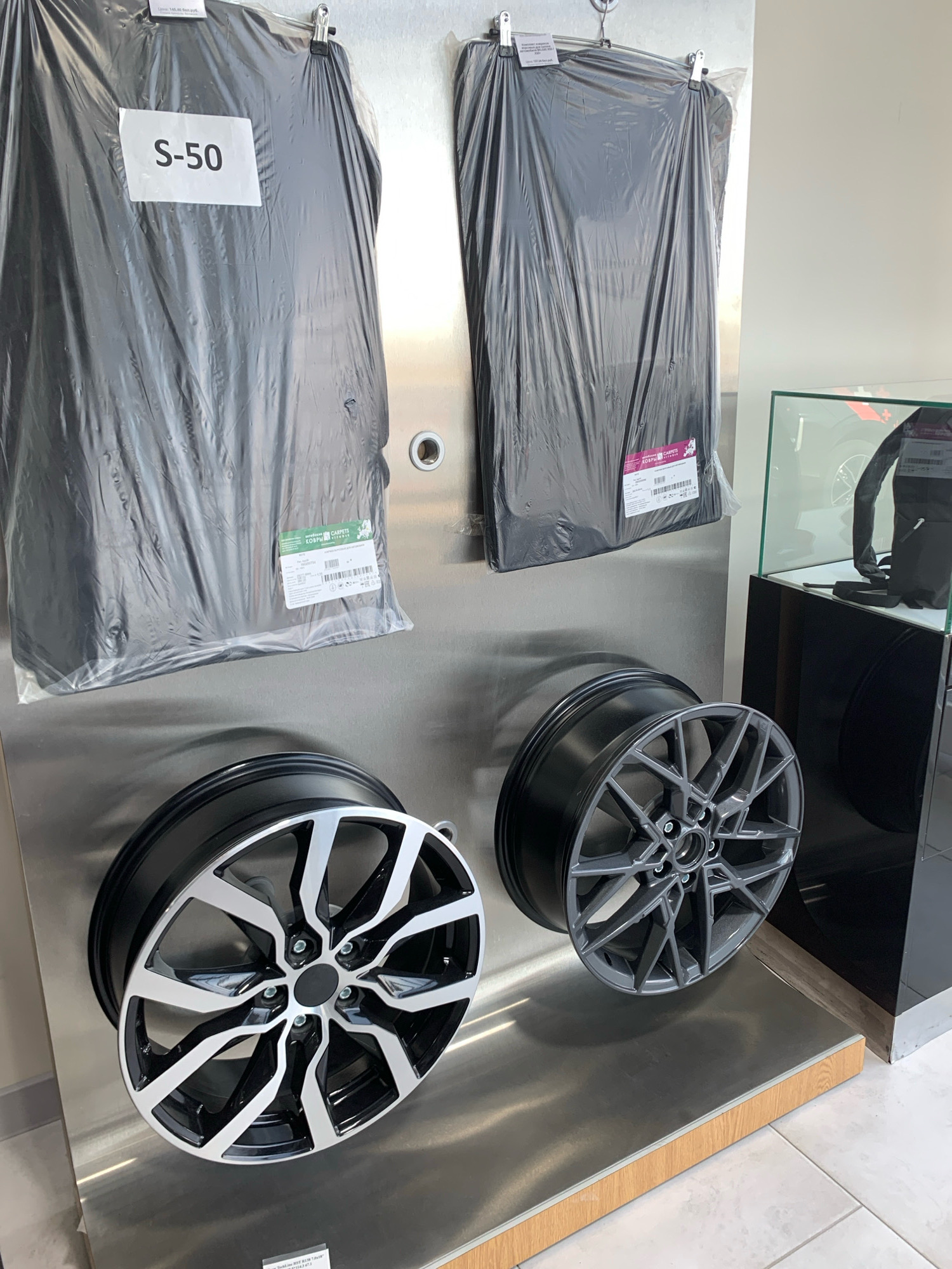 The image shows a display of car accessories in what appears to be a car dealership or parts store. In the upper portion of the image, two large black car floor mats are hung vertically, each enclosed in clear plastic. The mat on the left has a white label that reads "S-50". The mat on the right has a pink label with text in a foreign language, and another white label with text.
In the lower portion of the image, two car wheels are displayed horizontally. The wheel on the left is a two-tone alloy wheel with black and silver spokes. The wheel on the right is a darker grey alloy wheel with a more intricate spoke design.
To the right of the display, there is a glass-enclosed cabinet. Inside the cabinet, a black backpack is visible, along with some other items that are not clearly discernible. The background consists of a metallic wall and a tiled floor.
This scene is located in the city of Чыжэвіцкі сельскі Савет, Belarus, suggesting it is a retail environment within this region. There are no people visible in the image, and the focus is entirely on the product display.