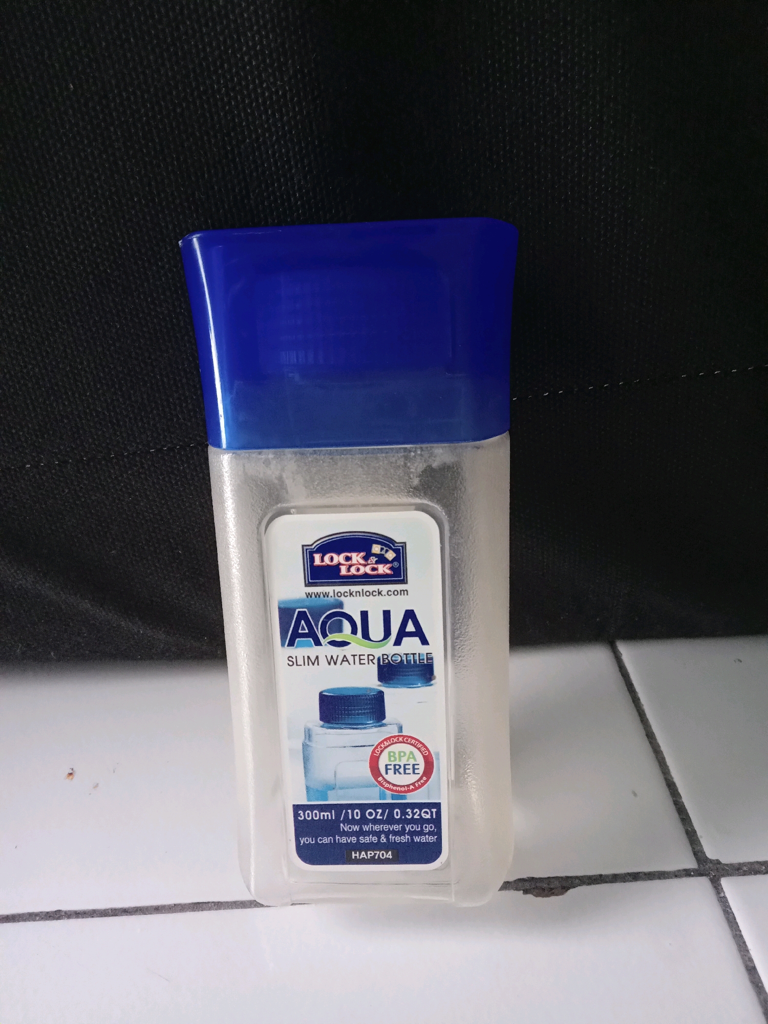 The image shows a transparent plastic water bottle with a blue cap and label. The label is white with blue and green text. At the top of the label is the "LOCK & LOCK" logo. Below that, it says "www.locknlock.com". Further down, it reads "AQUA SLIM WATER BOTTLE". In a circular red seal, it says "BPA FREE" and "Bisphenol-A Free". The bottom of the label indicates "300ml / 10 OZ / 0.32QT" and "Now wherever you go, you can have safe & fresh water". Below this is the model number "HAP704". The bottle is standing upright on a tiled surface. The background is a dark, textured fabric. This scene is not indicative of any specific location within the city of Bangah, Indonesia, as no identifiable landmarks or context clues are present.