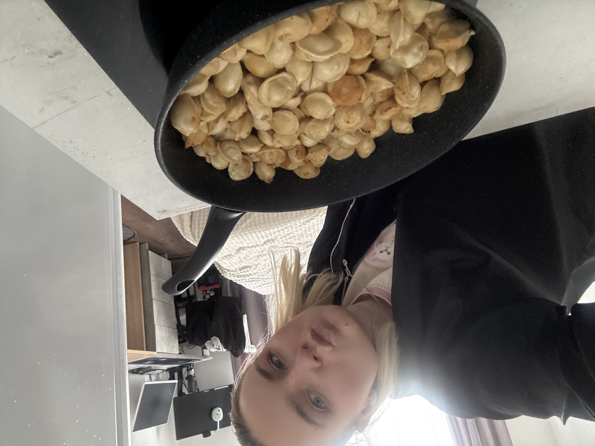 A person is holding a frying pan full of fried dumplings in a kitchen. The person has blonde hair and is wearing a black jacket over a light-colored top with a pink trim. The frying pan is black and contains many small, fried dumplings. The kitchen has gray countertops and walls. In the background, there is a desk with a computer monitor, and a bed can be seen behind the person. Based on the prompt, the location is Vsevolozhsk, Russia.