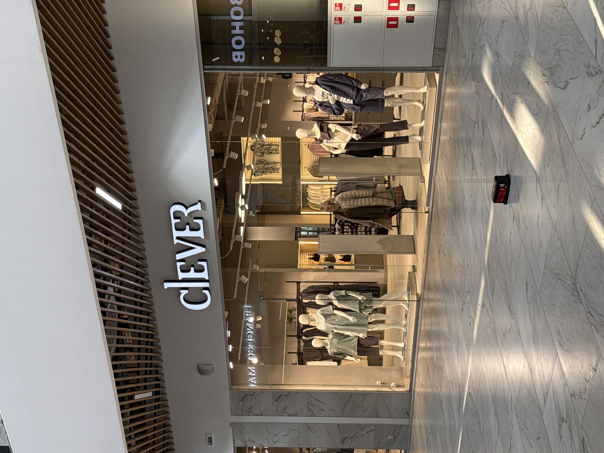 Here's a detailed description of the image:
**Setting:** The image captures the storefront of a clothing store named "CLEVER." It appears to be located inside a shopping mall or similar indoor retail complex, based on the polished marble or tile flooring and the presence of other stores or services nearby. The location is in Izhevsk, Russia, as "Уксобнов" is written in Russian on the window.
**Storefront:** The store's facade is primarily glass, providing a view into the display area. Several mannequins are dressed in various outfits, showcasing the store's clothing line.
**Objects:**
* **Mannequins:** Multiple mannequins display a range of clothing items, including tops, bottoms, and outerwear.
* **Clothing:** Various garments are visible on the mannequins, representing different styles and collections.
* **Signage:** The store's name, "CLEVER," is prominently displayed in a vertically oriented sign.
* **Fire Safety Equipment:** A small, red fire safety equipment or sign is situated on the floor near the storefront.
**Environment:** The environment is clean and well-lit. The floor has a polished appearance, and there are indications of other retail units in the vicinity.