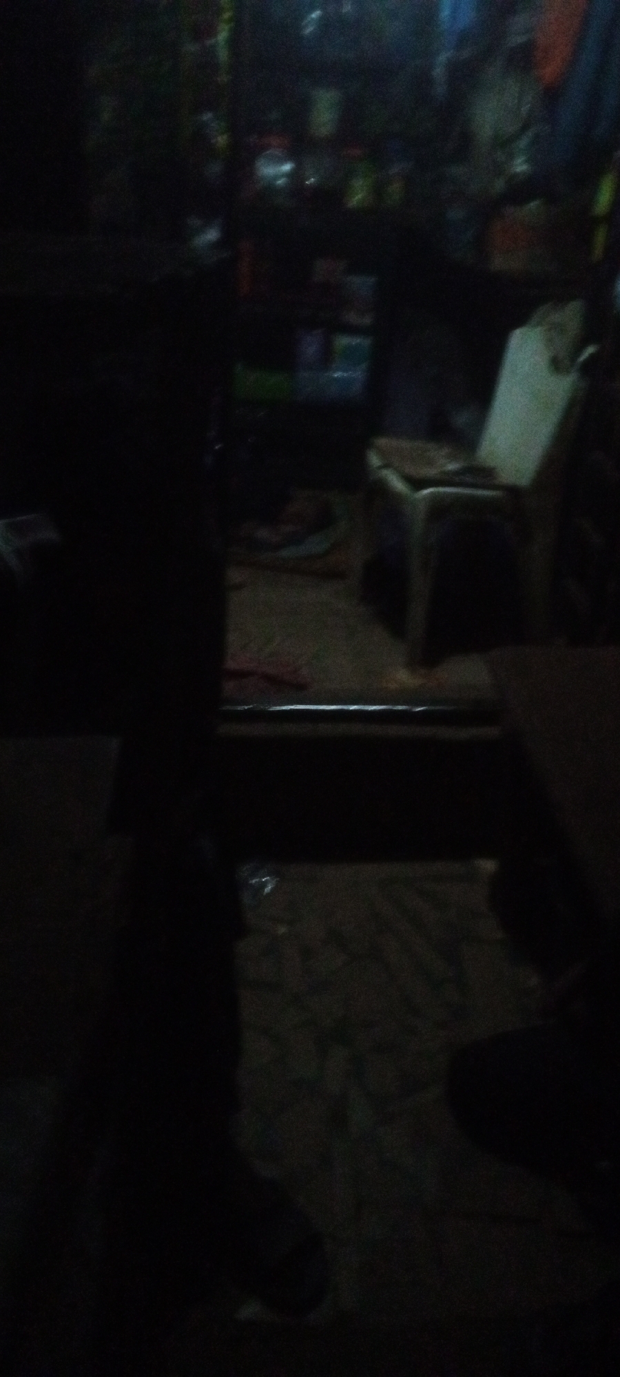 The image appears to be taken inside a dimly lit shop or room in Sabon Tasha, Nigeria. A person stands in the foreground, their features obscured by the darkness. Behind them, a metal barrier is visible, leading into the interior. A chair sits near the back, with shelves stocked with various items in the background. The floor has a patterned covering. The overall environment seems cluttered and dimly illuminated.
