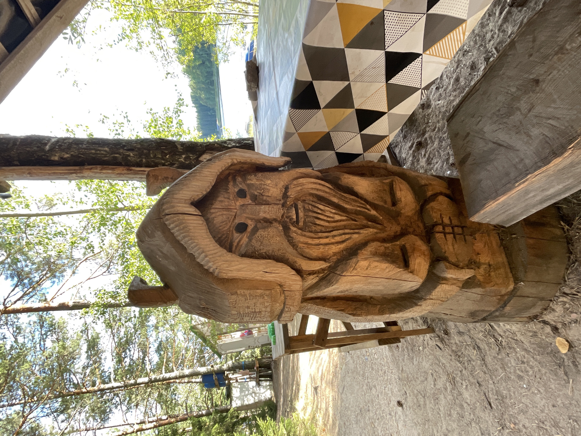 This image depicts a rustic outdoor scene, likely in Beryozovo, Russia, given the provided context, though the specific location within the city is not discernable from the visual information alone. In the foreground, a large, intricately carved wooden sculpture of a man with a beard and a hooded garment stands prominently. The sculpture appears to be made from a single piece of wood and exhibits visible tool marks. To the right of the sculpture, a picnic table is covered with a geometrically patterned tablecloth featuring triangles in shades of grey, yellow, and black. A stone rests on the tablecloth. Behind the table, a body of water, possibly a lake, is visible, bordered by a dense forest of coniferous and deciduous trees under a bright, sunny sky. In the background, part of a wooden structure, possibly a gazebo or shelter, is visible, along with trees and a glimpse of what might be an adventure park with platforms and blue barrels. The ground is sandy and covered with dried leaves and pine needles. No people are visible in the frame. The overall atmosphere is serene and suggests a recreational area.
