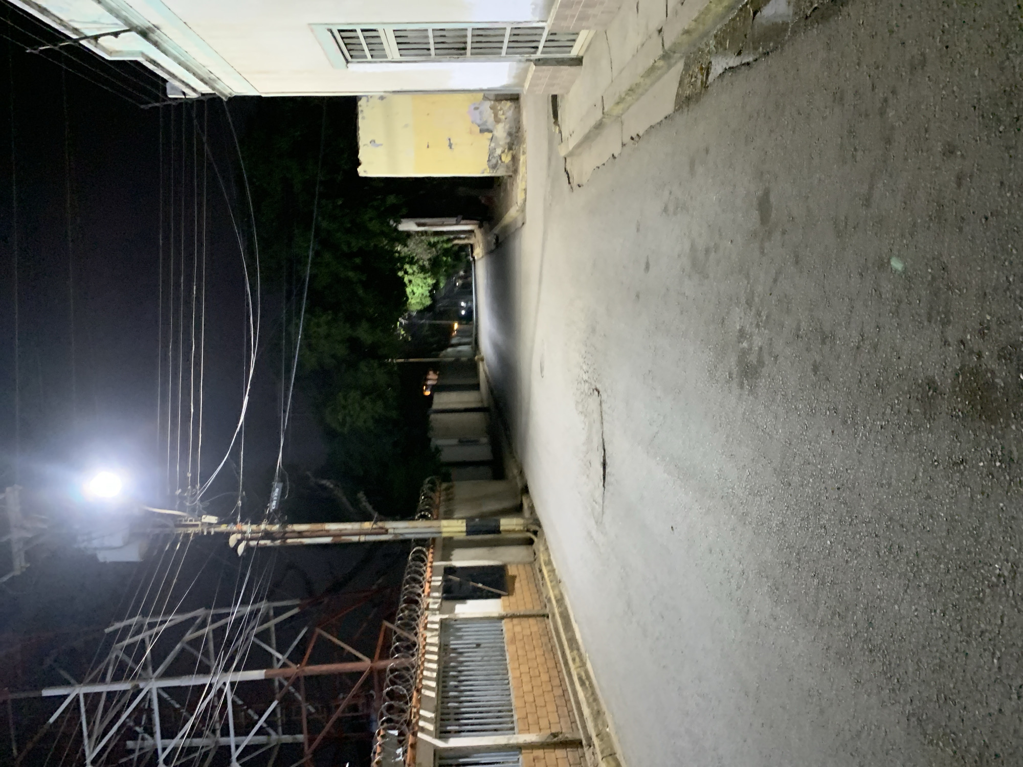 This is a nighttime photograph of a deserted street or alley in Santa Ana, Venezuela, taken from a slightly elevated perspective looking down the thoroughfare. The scene is illuminated by artificial light sources, primarily a bright streetlamp on the left.
On the left side of the image, a utility pole is prominent, featuring a bright, glaring streetlamp and a dense tangle of overhead power lines and cables extending across the frame. Below these wires, a large, intricate metal lattice structure, possibly part of a communication tower or an industrial framework, is visible. Along the top of a low wall or fence bordering this structure, coils of barbed wire are present.
The right side of the image features a paved road surface, appearing gray and somewhat weathered with visible cracks and texture, especially in the foreground. A raised concrete sidewalk or curb runs alongside the road. Adjacent to the sidewalk are buildings. The closest building has a light-colored facade with a barred window and sections of peeling yellow paint, revealing damaged underlying layers. Further down the street, additional building structures can be discerned, some featuring corrugated metal cladding and brickwork. A small, light green object rests on the asphalt surface in the mid-ground.
The street recedes into the darkness, with dark foliage from trees or bushes visible in the background and faint, distant lights suggesting other structures or activity further along. No people or vehicles are present, lending a quiet and somewhat desolate atmosphere to the scene. There is no discernible text visible in the image.