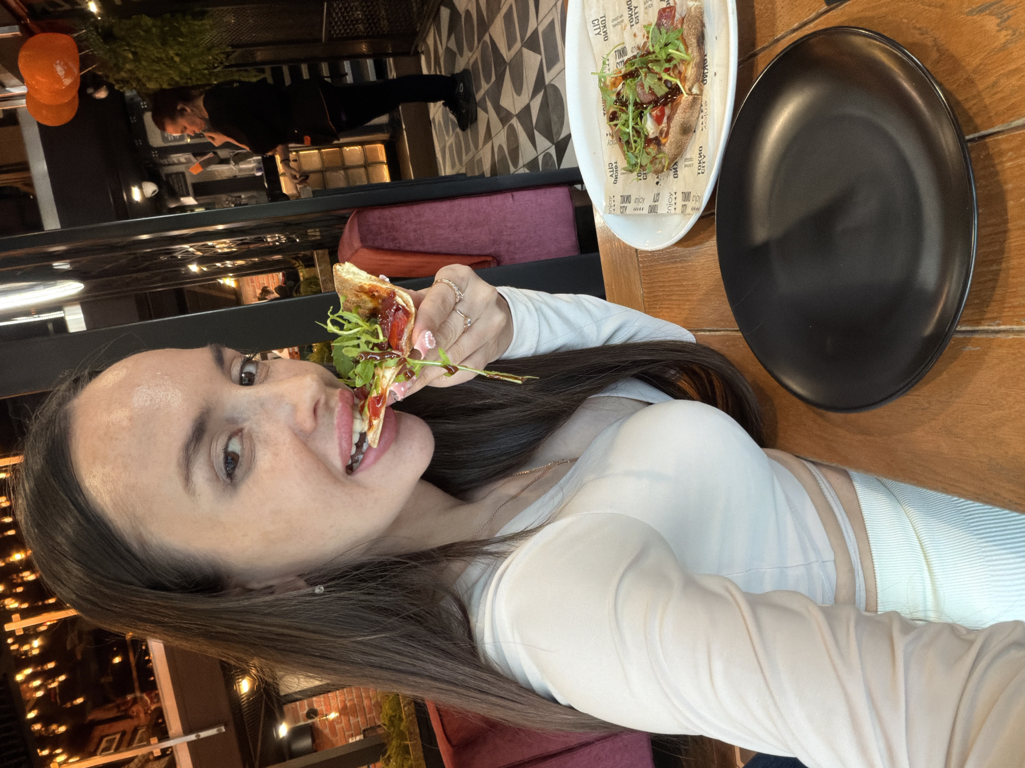 A woman with long, dark brown hair and light skin is depicted from the chest up, looking directly at the camera while biting into a piece of flatbread or pizza. She wears a light beige or white long-sleeved crop top that reveals a sliver of her midriff, paired with matching ribbed pants or skirt. A thin chain necklace with a small pendant is visible, along with rings on her right hand, which holds the food. Her nails are manicured in pink with white dots. The food she is eating, and another piece on a white plate to her right, is topped with arugula and a dark sauce, consistent with a flatbread or thin-crust pizza. The paper liner beneath the food on the white plate clearly displays "TOKIO CITY" branding, indicating the location is a Tokio City restaurant, a chain known to operate in Saint Petersburg, Russia.
The scene is an indoor restaurant with a warm ambiance. The woman sits at a wooden table with a black empty plate to her left. Behind her, a red velvet upholstered banquette is visible. The background on the left features exposed brickwork and warm string lights, suggesting an upper level or mezzanine. On the right side of the background, a person in dark clothing, appearing to be a staff member, stands near a counter or bar area. This area includes metallic elements, glass blocks, and overhead orange spherical decorations (possibly lamps or balloons). The floor in the background features a patterned black and white tile design.