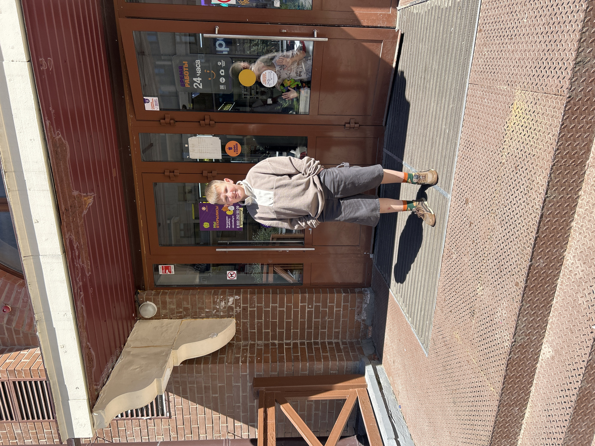 The image shows a young boy standing in front of a store entrance. He is wearing a beige sweatshirt, dark gray shorts, orange and green socks, and brown hiking shoes. The store has double glass doors with signs in Russian. One sign says "WE ARE OPEN" and another says "24 HOURS". The building is made of red brick and has a covered entryway with a metal grate and a diamond plate ramp. There are shadows on the ground, indicating a sunny day. The boy is looking at the camera with a slight smile. The location is identified as Khimki, Russia.