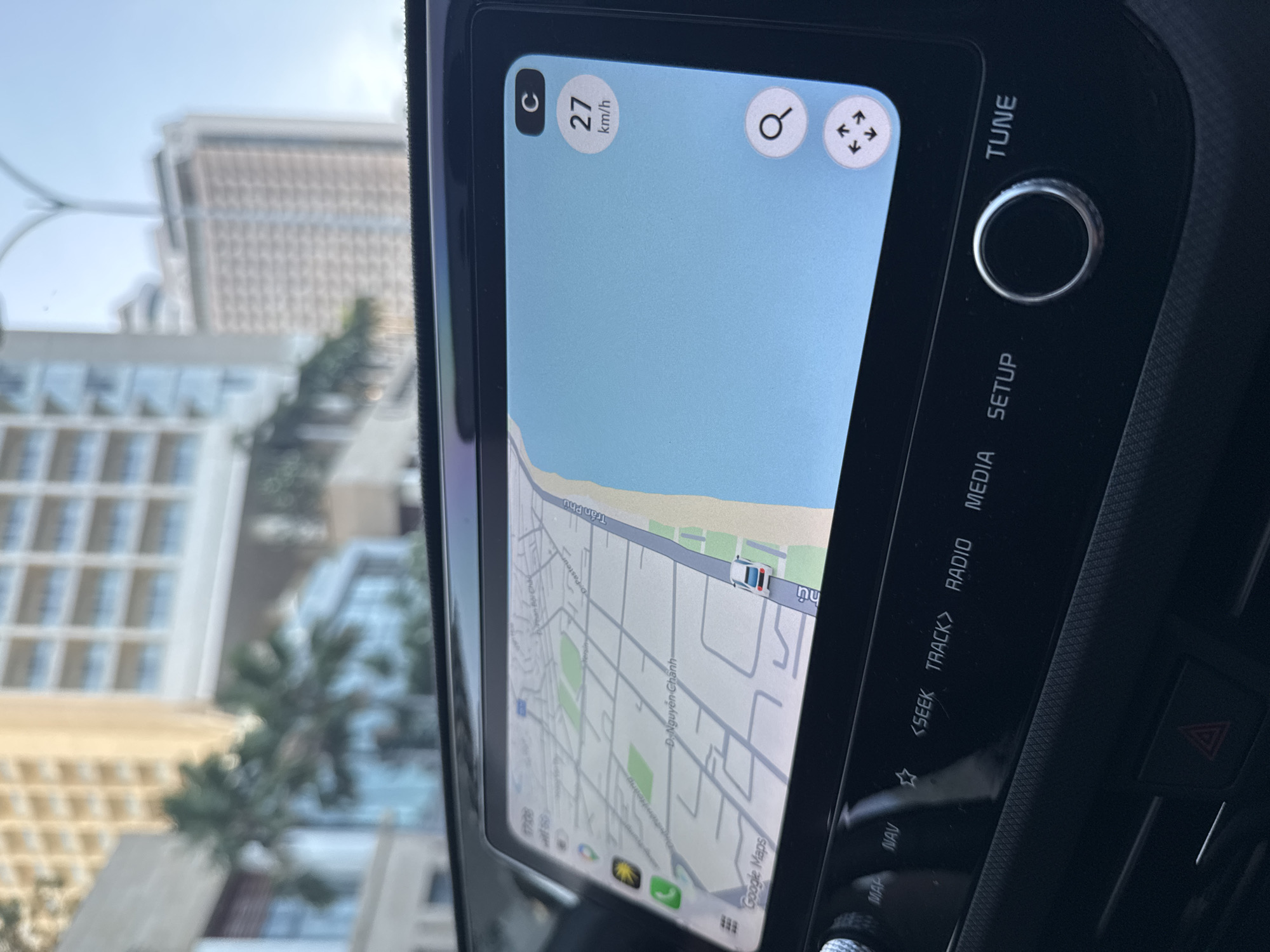 The image depicts the interior of a vehicle, specifically the dashboard and infotainment system displaying a navigation map, with an out-of-focus view of an urban coastal environment through the windshield.
In the foreground, the car's infotainment screen shows Google Maps with a current speed of 27 km/h. The map illustrates a coastal road, identified as Trần Phú, running parallel to a body of water (the sea) with a beach strip in between. Other visible street names on the map include D. Nguyễn Chánh, D. Pasteur, and Phan Bội Châu, all located within Nha Trang, Vietnam, specifically pointing to Lộc Thọ Ward due to the intersection of D. Nguyễn Chánh and Trần Phú Street. The screen also displays an Apple CarPlay interface with the time 17:09. Below the screen, physical buttons for car functions are visible, including "MAP," "NAV," "SEEK," "TRACK," "RADIO," "MEDIA," and "SETUP," along with a "TUNE" knob and a hazard light button.
The background, visible through the windshield, shows several modern high-rise buildings and palm trees under bright daylight, characteristic of a developed resort city. No specific individuals are discernible.