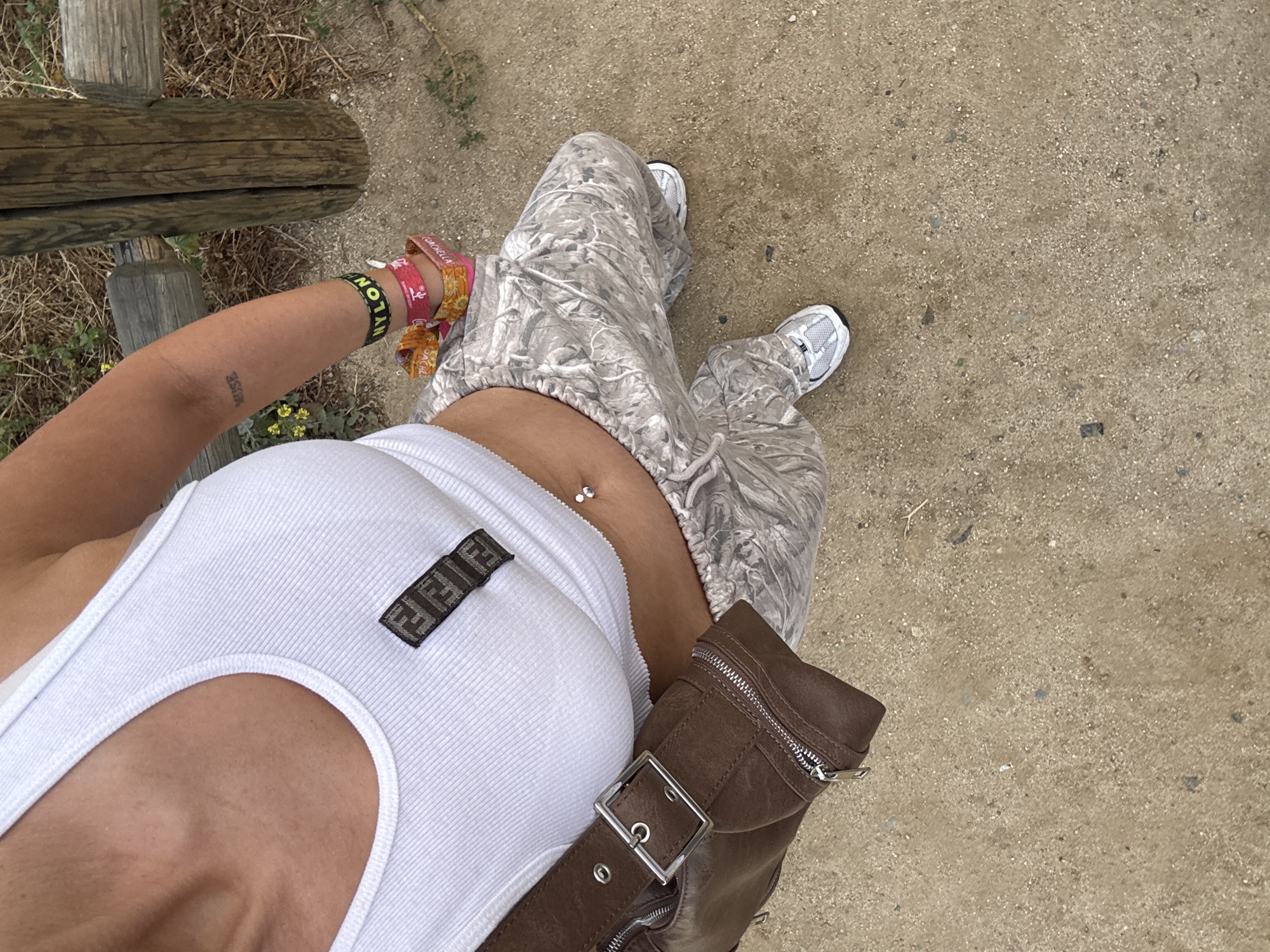 A top-down medium shot depicts a person standing on a tan, sandy surface in an outdoor setting. The person wears a white ribbed tank top with a Fendi logo patch, camouflage print pants, and white sneakers. A brown leather fanny pack is strapped across the body. The person has multiple colorful wristbands on their left wrist and a tattoo on their left arm. A wooden structure and sparse vegetation are visible in the background. The location is suggested to be in Los Angeles, United States of America.