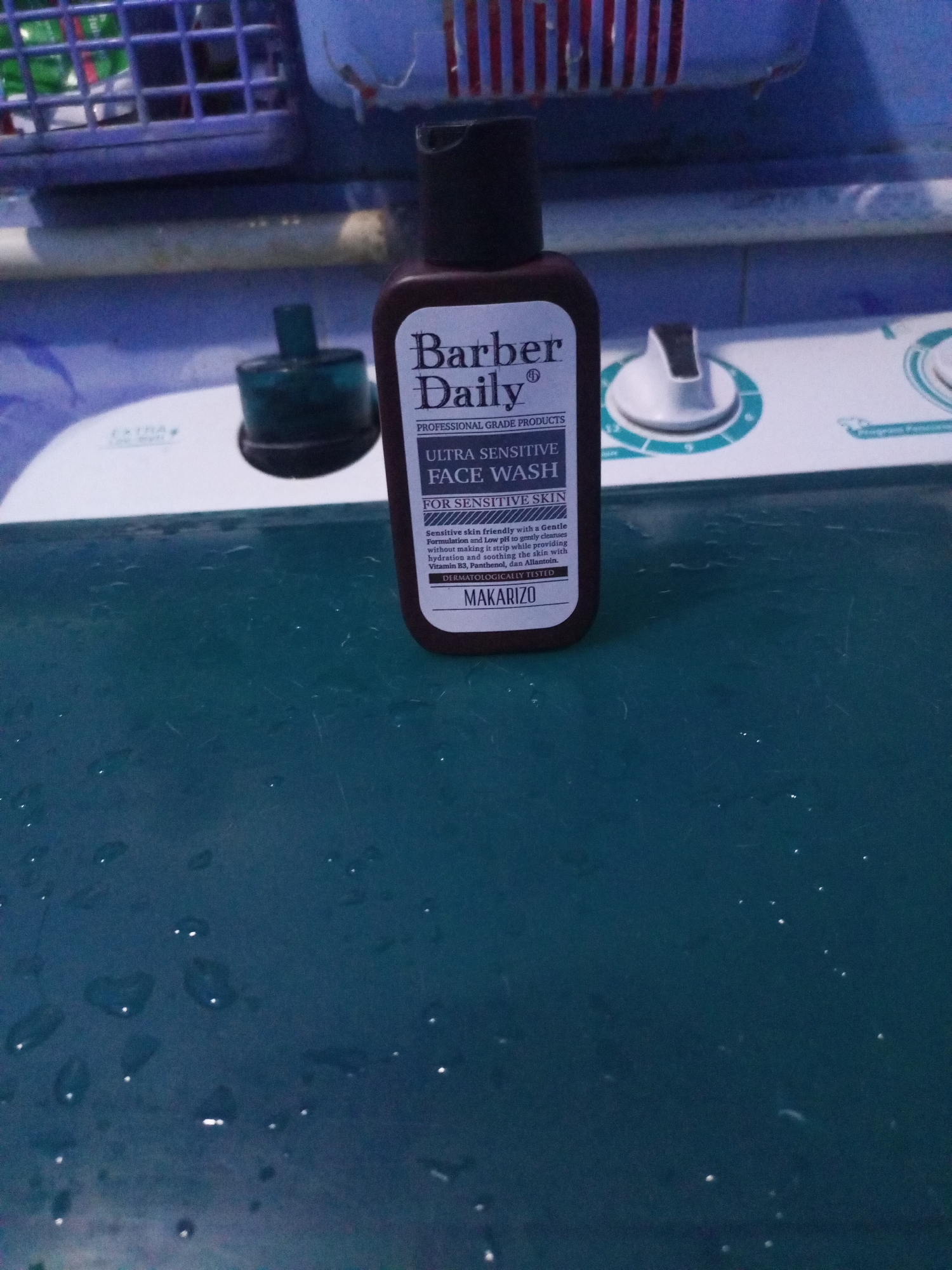 A close-up shot focuses on a brown plastic bottle of "Barber Daily" face wash, positioned on a dark teal, wet surface. The bottle is upright and centered in the frame, with its label clearly visible. The label features the text "Barber Daily" in large, bold letters, followed by "PROFESSIONAL GRADE PRODUCTS" and "ULTRA SENSITIVE FACE WASH." Smaller text details the product's formulation for sensitive skin, mentioning gentle cleansing, low pH, hydration, soothing properties, and ingredients like Vitamin B3, Panthenol, and Allantoin. Below this, "DERMATOLOGICALLY TESTED" and "MAKARIZO" are visible.
In the blurred background, a light-colored washing machine or similar appliance is partially visible, with control knobs and a ribbed blue top. To the left of the bottle, a dark blue cap, possibly from another product, sits on the same surface. The teal surface appears to be a countertop or table, with numerous small water droplets scattered across it, reflecting the light. The overall impression is one of a bathroom or laundry area. The image is likely taken in a residential setting in Cidahu, Indonesia, given the context of a daily personal care product being used in such an environment.