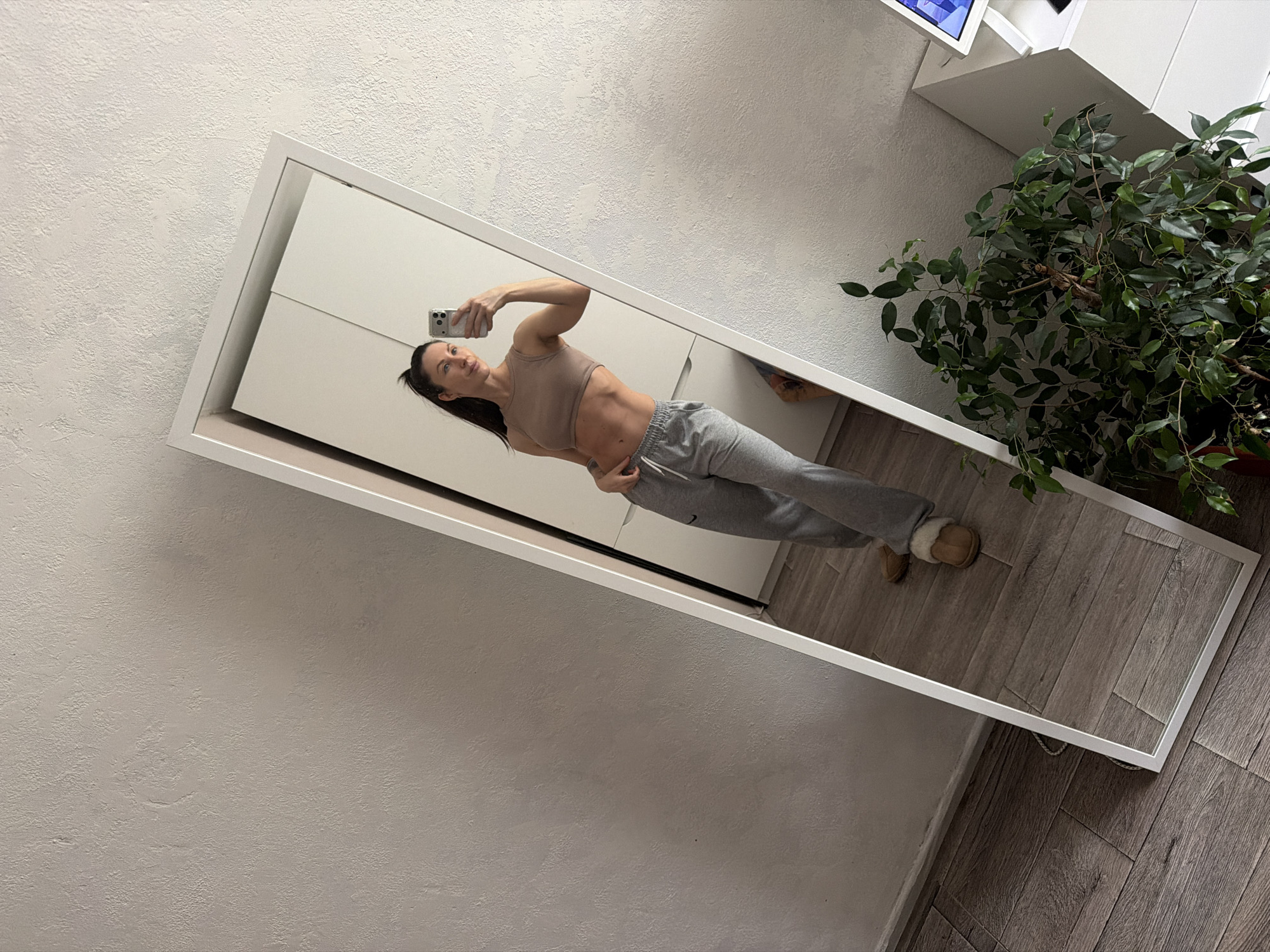 A woman is taking a mirror selfie inside a building. The mirror is leaning against a wall with white cabinetry and a potted plant visible. The woman is wearing a sports bra and sweatpants. The flooring is wooden. The building appears to be located in Perm, Russia, based on prior information.
