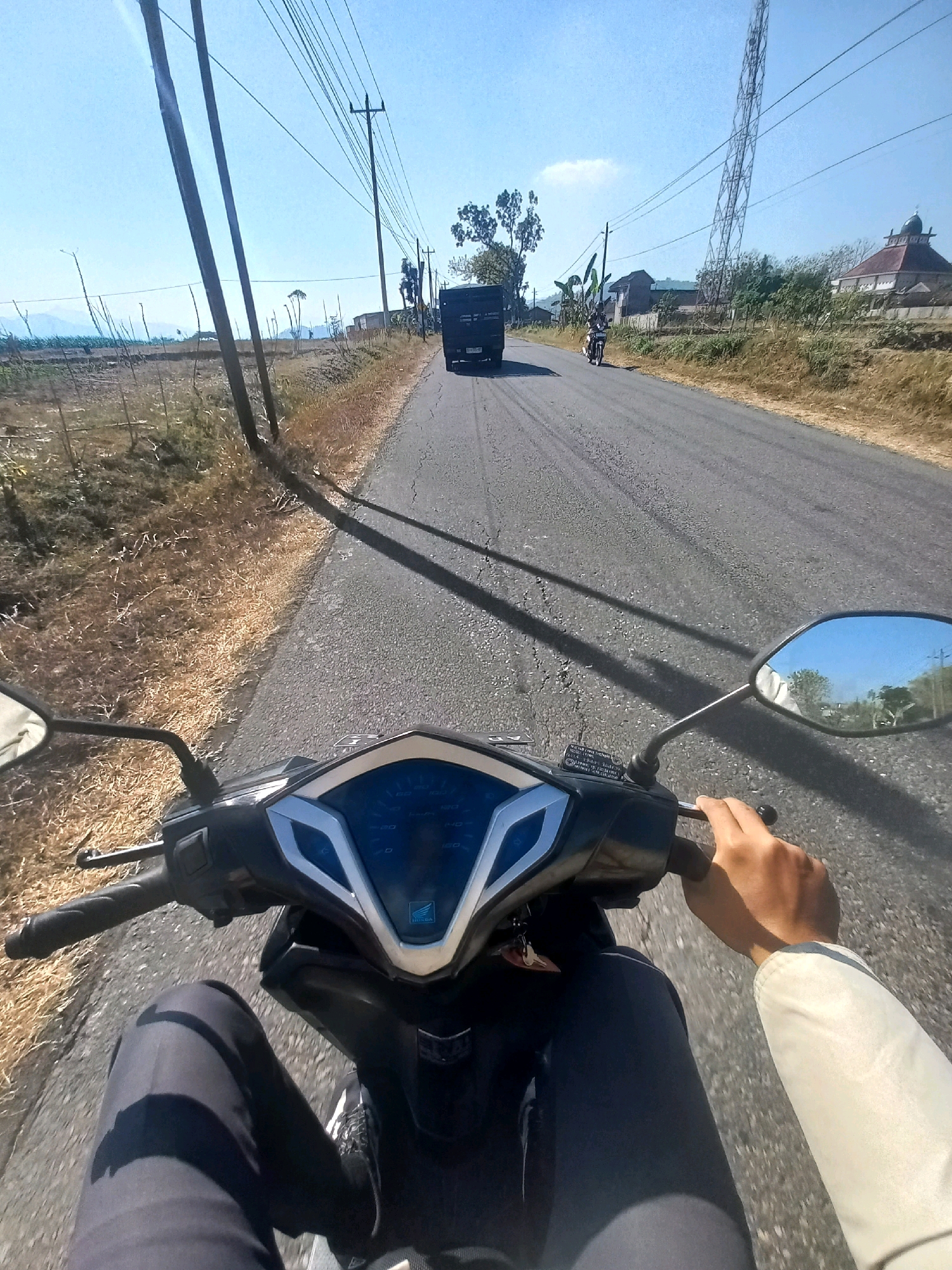 The image shows a first-person perspective of someone riding a motorcycle on a paved road in Baturetno, Indonesia. The road is flanked by dry, grassy fields and scattered trees. In the distance, there are buildings, including what appears to be a mosque with a domed roof. Utility poles and power lines run along the side of the road, and a cell tower is visible in the background. A black truck is ahead on the road, and another motorcycle is further in the distance. The sky is clear and blue. The rider's hands are on the handlebars, and their legs are visible in dark pants.