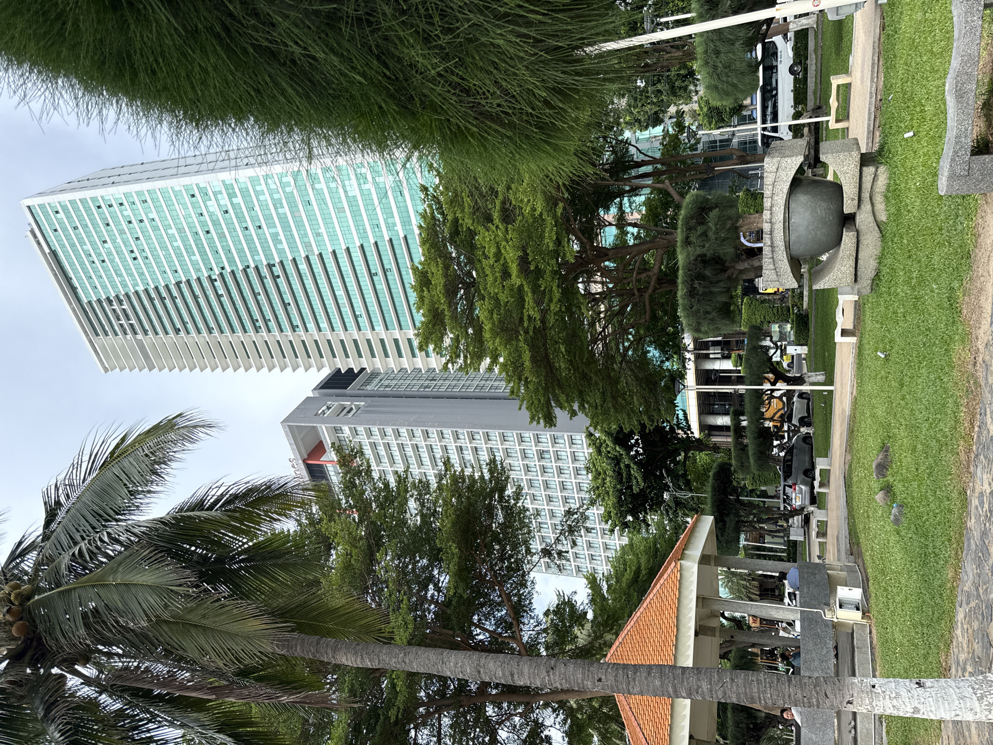 The image depicts a street scene in Nha Trang, Vietnam, with a prominent view of the vast, modern architectural landscape juxtaposed with lush greenery and a gazebo. In the foreground, a mature palm tree stands tall, its fronds reaching towards the overcast sky. To its left, a gazebo with an orange-tiled roof offers a shaded respite, with several people seated inside.
The middle ground is occupied by manicured green lawns, a unique stone sculpture resembling an eye or a vessel, and several benches. Beyond the greenery, a grey sedan and a white van are visible, suggesting a public road adjacent to the park.
The background is dominated by two imposing skyscrapers. The one on the left is a lighter-colored building with numerous balconies, while the taller, dominant structure on the right features a striking facade of turquoise glass windows and white horizontal accents, indicative of modern high-rise architecture. The overall impression is one of urban development coexisting with natural elements within a city.
