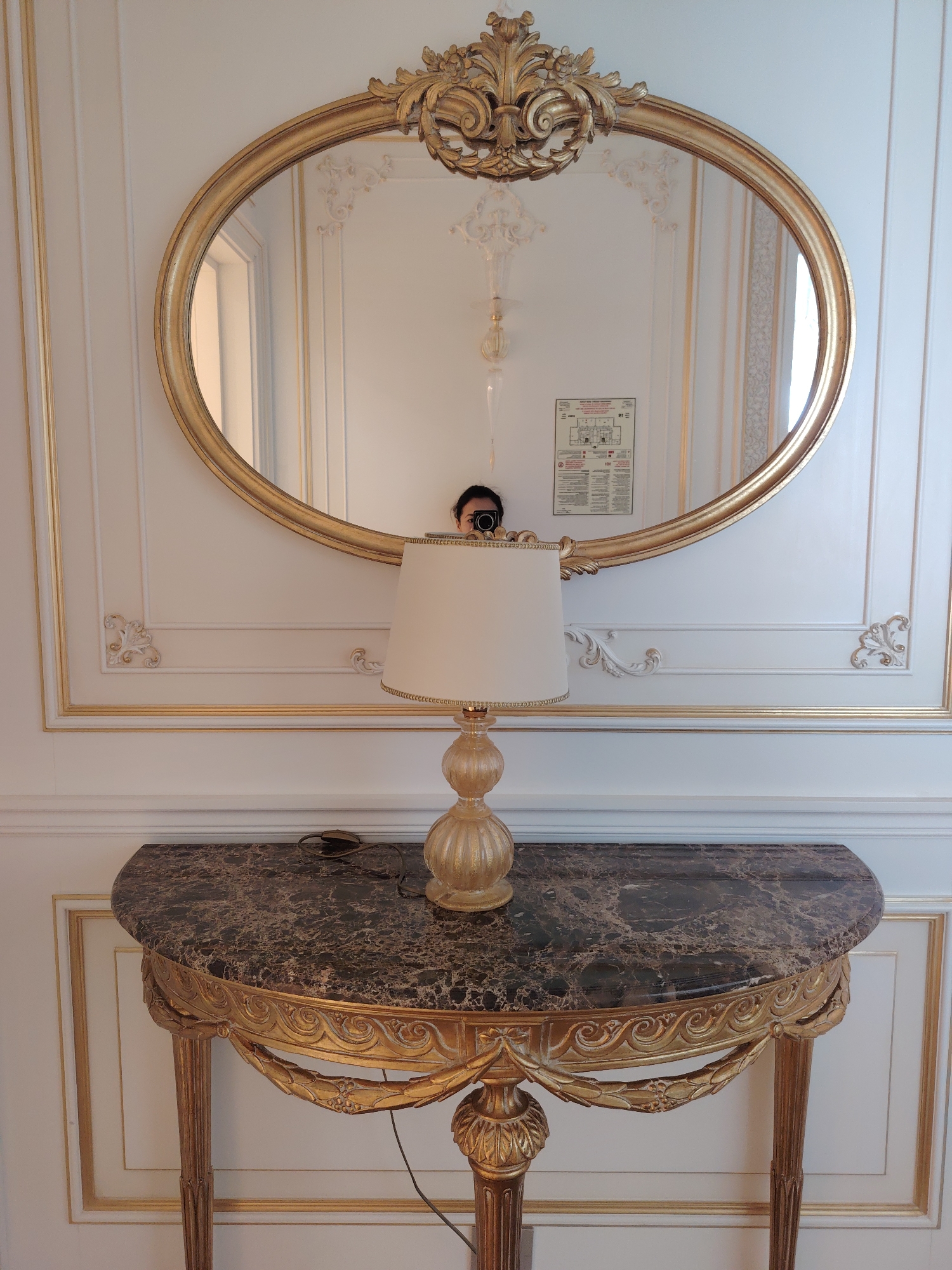 The image depicts an interior scene featuring a console table with a marble top and ornate gold detailing, a lamp on the table, and a large oval mirror with an elaborate gold frame hanging on a white wall. The wall itself has decorative gold trim and paneling. In the reflection of the mirror, a person holding a camera is visible, taking the photograph. A fire safety notice is also visible on the wall in the mirror's reflection. The style of the decor suggests a classical or baroque influence. The image is a photograph taken from a medium distance, capturing the central arrangement of furniture and the mirror.