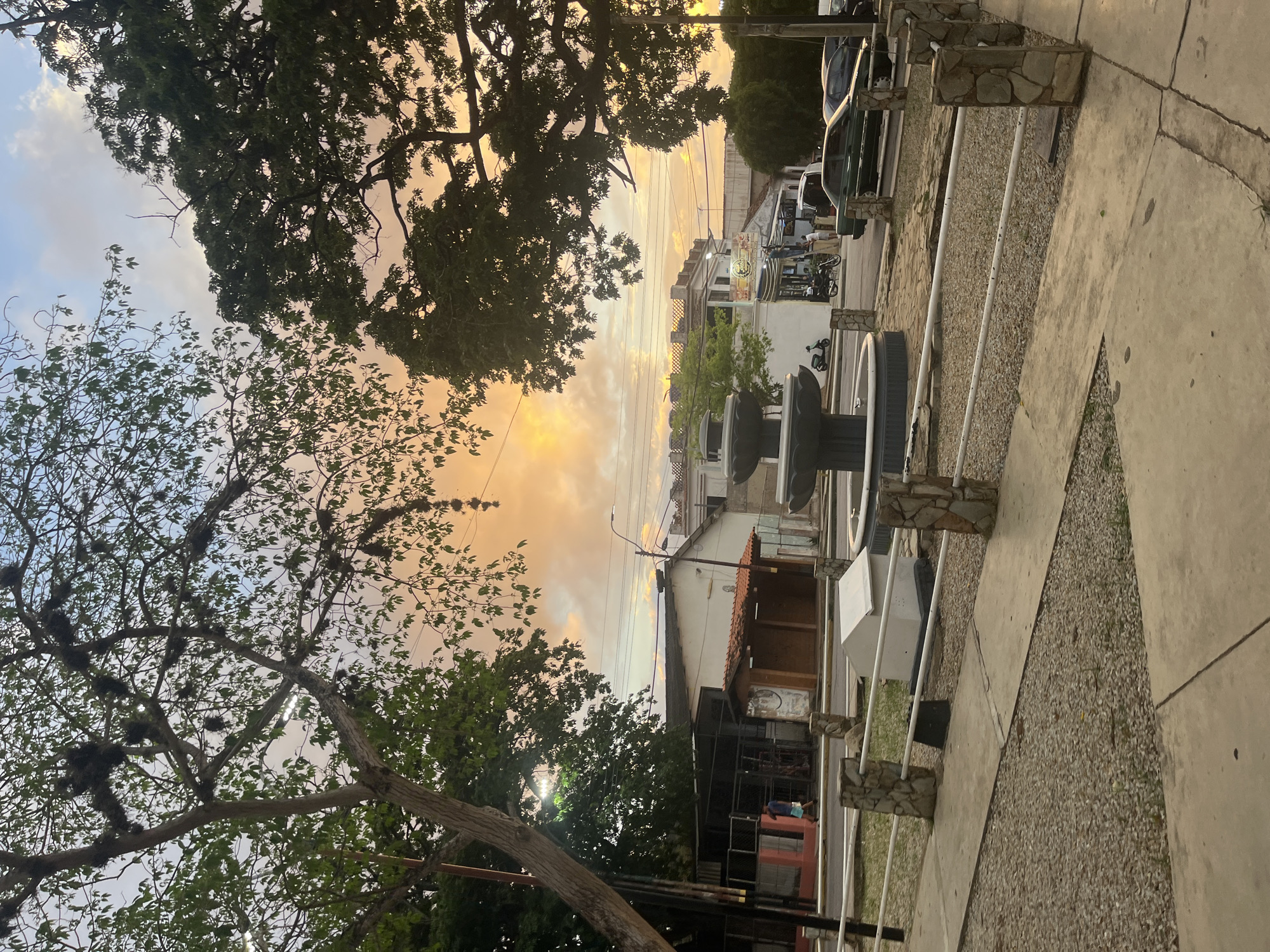 The image depicts an outdoor scene at dusk in what appears to be a public square in Santa Ana, Venezuela. The sky is filled with warm, orange and yellow hues, indicating sunset. In the foreground, a concrete pathway is bordered by a low fence made of white metal bars supported by stone pillars. Beyond the fence, a gravel-covered area is visible, with more pathways and a white, box-like structure.
Dominating the middle ground is a multi-tiered fountain, dark in color, with water not actively flowing. Behind the fountain, buildings line a street. One building has a white facade and an orange-tiled roof, possibly a small shop or kiosk. Further down the street, other buildings with awnings and signage are visible. Several vehicles, including a dark green sedan and a black car, are parked or driving on the street. A motorcycle and a scooter are also present.
Two trees with lush green foliage frame the scene, with one on the left and another on the right. The tree on the left has some aerial roots or epiphytes hanging from its branches. In the background, utility poles and wires crisscross the sky. A single pedestrian, a man wearing a blue shirt and shorts, is walking on the sidewalk in front of a building with a fenced-in area. The overall impression is a quiet, urban twilight scene.