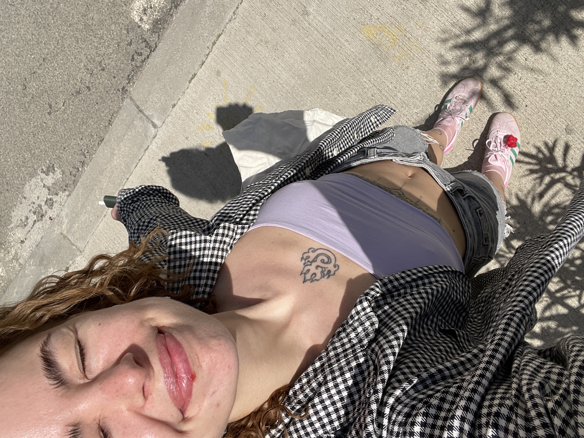 A young person lies on a concrete sidewalk in direct sunlight. The location is not discernible as Boreti, Montenegro, from the image alone. The individual has curly, reddish-brown hair, and their eyes are closed. Their lips appear chapped or injured. They are wearing a light purple tank top, grey denim shorts, and a black and white houndstooth jacket. A tattoo is visible on their chest. Pink sneakers with a decorative red flower adorn their feet. A white tote bag lies beside them on the sidewalk, and they are holding a dark object in their left hand. Shadows from foliage are cast on the ground.