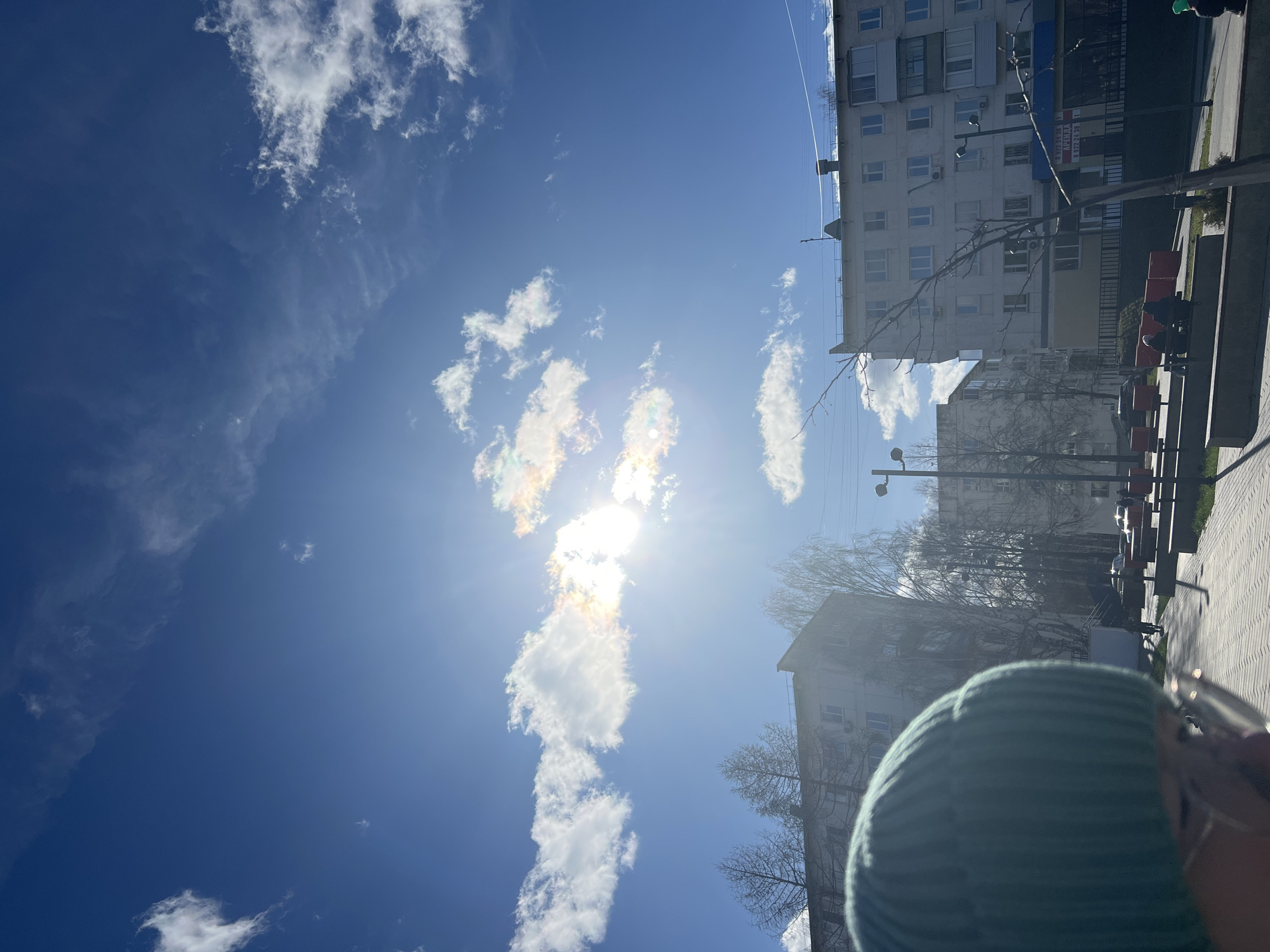 The image shows a view of the sky and a street in Chaikovsky, Russia. The sky is bright blue with scattered white clouds, some displaying iridescence due to sunlight diffraction. Below the sky, there are buildings. A tall, apartment-style building with white exterior walls and multiple windows is visible. Streetlights line the walkway. A person wearing a blue-green striped beanie hat is partially visible at the bottom of the frame. Pedestrians are visible on the benches. The image is rotated ninety degrees.