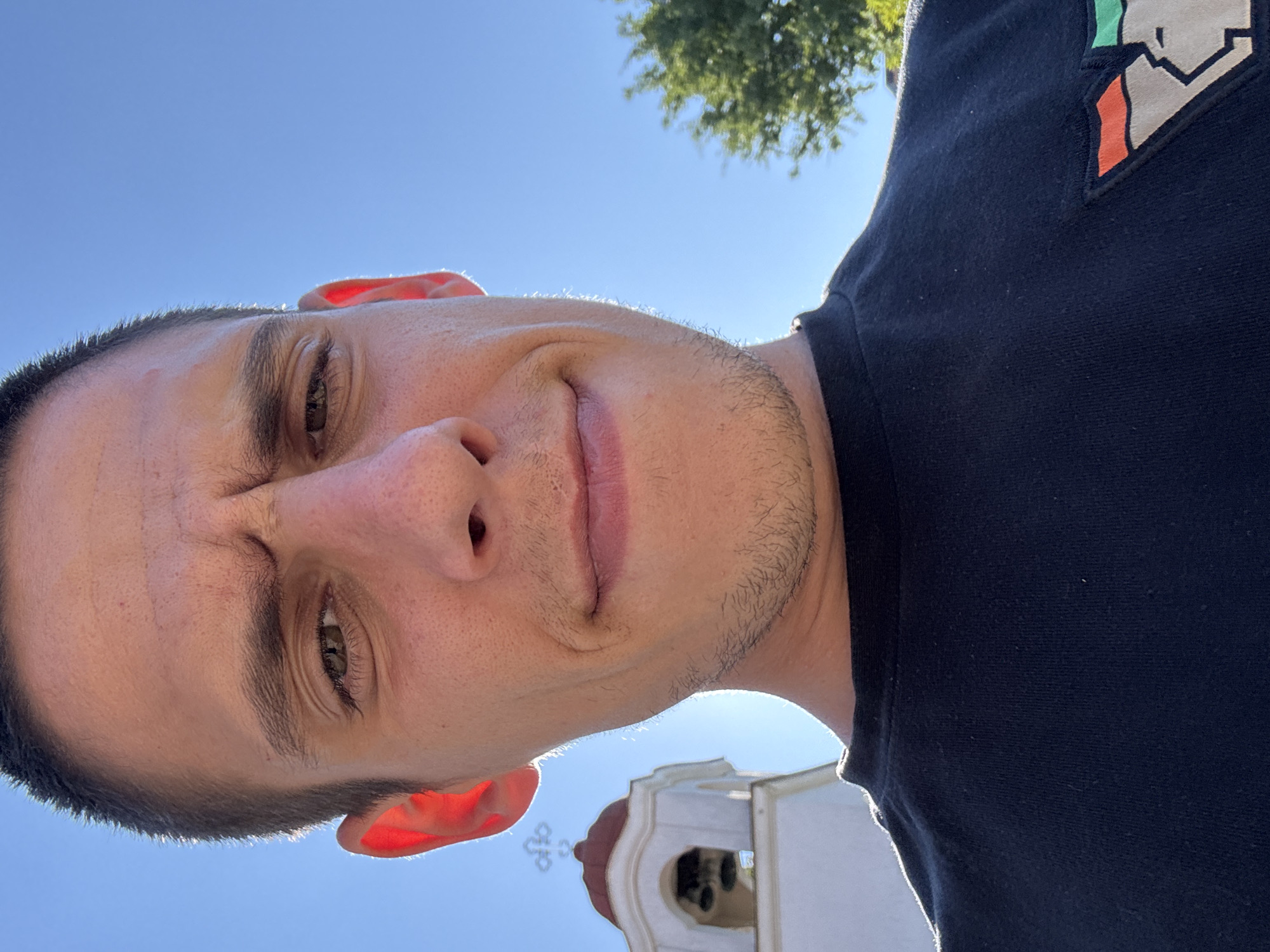A close-up, low-angle selfie of a young man with short dark hair, taken outdoors on a sunny day. He is wearing a black t-shirt with a patch on the right side of his chest featuring a stylized red, green, and grey design. The man has a slight smirk and is looking directly at the camera. His left ear is brightly illuminated by the sun. In the background, a white church bell tower with a red dome is visible, along with green foliage and a clear blue sky. The overall impression is of a casual outdoor setting, possibly a public square or park in Sofia, Bulgaria, given the architectural style of the bell tower.