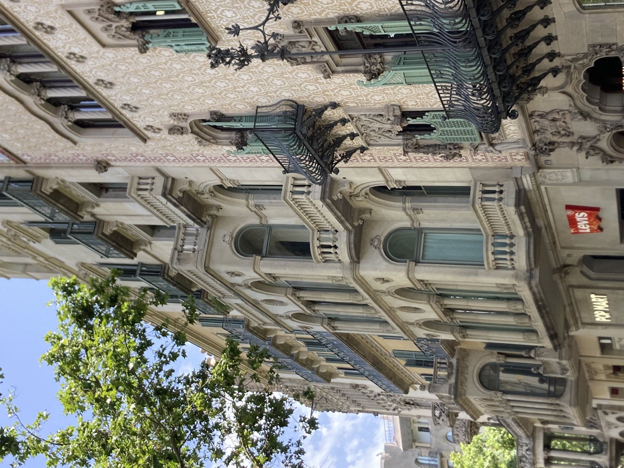 This is a vertical, medium shot of the facade of an ornate building in Barcelona, Spain. The architecture is very detailed, with intricate carvings, decorative balconies with wrought iron railings, arched windows, and patterned stonework. The building appears to be several stories high, with each level featuring a different style of ornamentation. A tree with lush green leaves is partially visible in the lower left, reaching up towards the sky, which is a bright blue with some white clouds. Sunlight illuminates the building, suggesting it is daytime. The overall scene conveys a sense of historic grandeur and urban beauty. Visible text includes "Levis" in red on a square sign, and "POP MART" on a vertical sign. The image is captured at an angle, giving a unique perspective on the building's complex design.