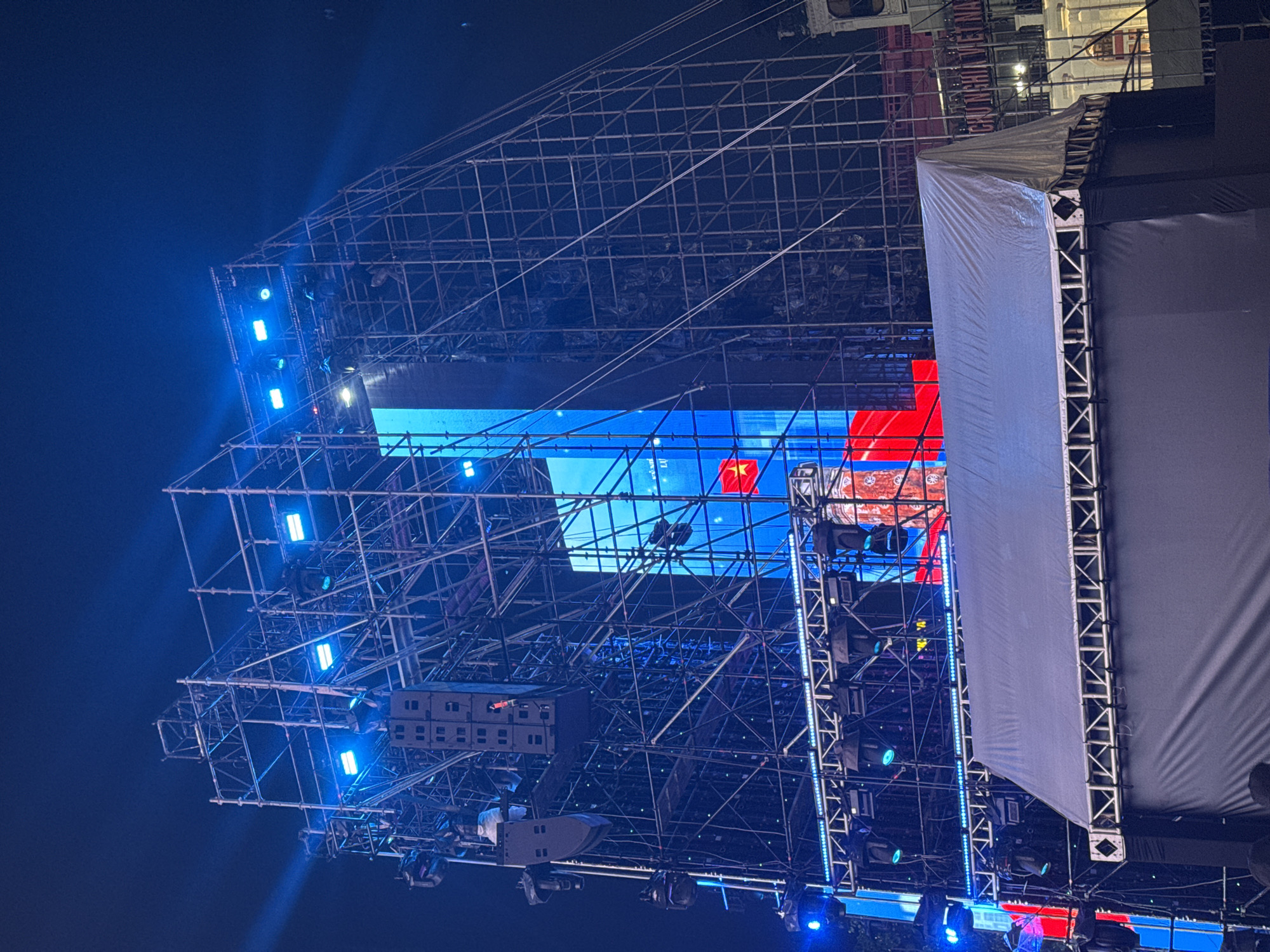 This image depicts a large outdoor stage setup at night, likely for a concert or public event. The stage is adorned with extensive scaffolding, speaker systems, and large LED screens displaying vibrant blue and red graphics, including a Vietnamese flag. Numerous lights are illuminating the scene, creating a dramatic effect. In the background, a building with illuminated signage that reads "CHÚNG NGHĨA VIỆT NAM" is visible, identifying the location as Hanoi, Vietnam. The scene appears to be in a public square or park, with a crowd of people faintly visible in the foreground. The overall atmosphere is festive and energetic, indicative of a significant cultural or political event.