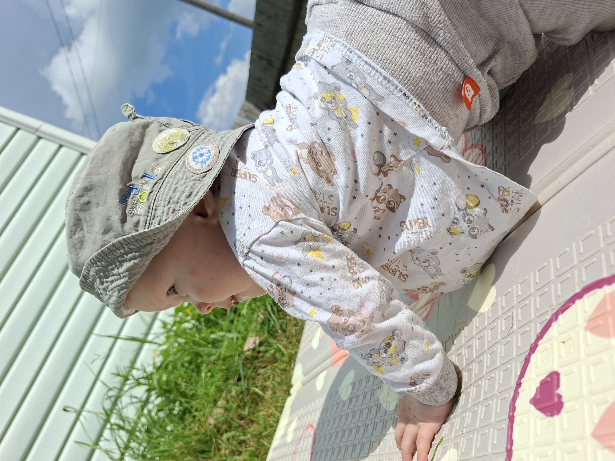 An infant wearing a grey bucket hat and a long-sleeved white shirt with a pattern of cartoon bears and the words "SUPER STAR" is on its stomach on a textured mat outdoors. The background shows a light green corrugated wall, green grass, and a bright blue sky with clouds. The infant is facing to the left, looking slightly upwards. The sun is casting shadows, indicating it is daytime.