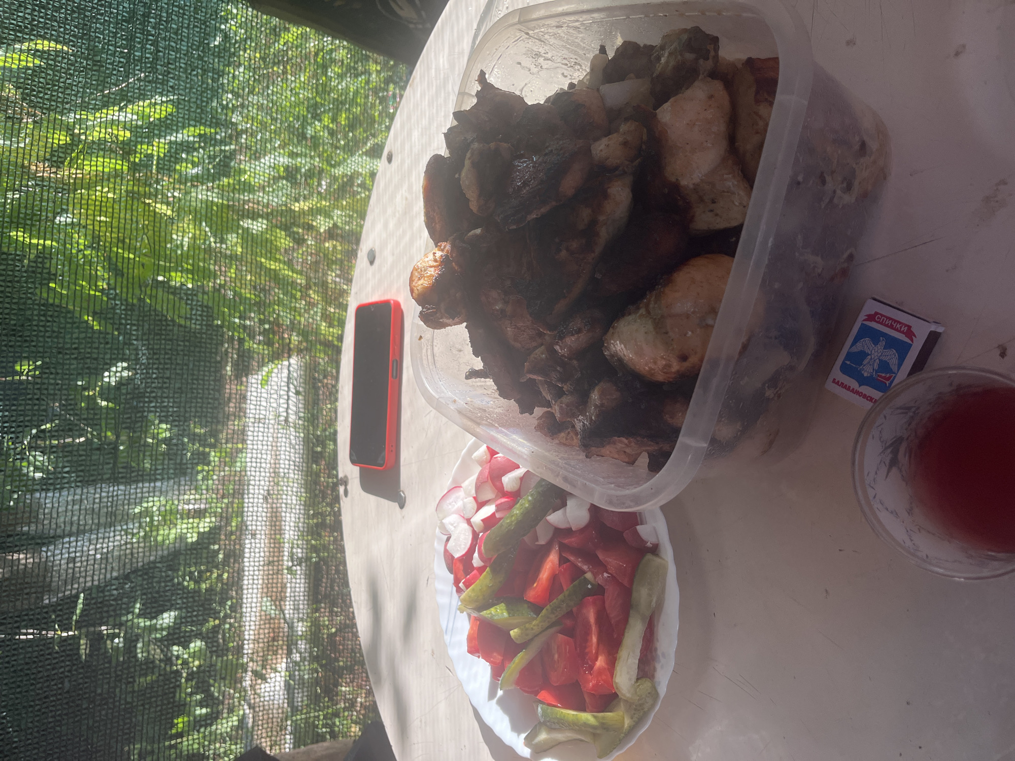In an outdoor setting, possibly Rakhinka, Russia, a table is set for a meal. A white plate holds a salad of chopped tomatoes, cucumbers, and radishes. Next to it, a clear plastic container is filled with grilled meat, appearing to be chicken or pork, with some pieces charred. A glass of red liquid, likely a beverage, sits in the foreground. A box of matches is placed near the glass. A red smartphone rests on the table in the background. The scene is partially shaded by a green mesh screen, through which sunlight filters, illuminating lush green foliage.