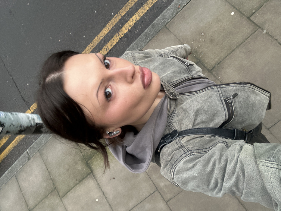 The image depicts a high-angle, selfie-style close-up of a female individual standing on a concrete pavement in the City of London, United Kingdom. Adjacent to the pavement is an asphalt road marked with double yellow lines. A light-colored utility pole is partially visible on the pavement to the upper right.
The female subject has dark hair parted centrally, brown eyes, and fair skin, with visible makeup including blush on her cheeks and defined eyebrows. She wears a grey hooded top layered beneath a distressed grey-green zippered jacket with snap button accents. A black strap, consistent with a shoulder bag, crosses her left shoulder. A silver-colored earbud is present in her left ear. She is looking directly into the camera with a neutral expression, her lips slightly parted in a pout. The environment indicates overcast daylight conditions.