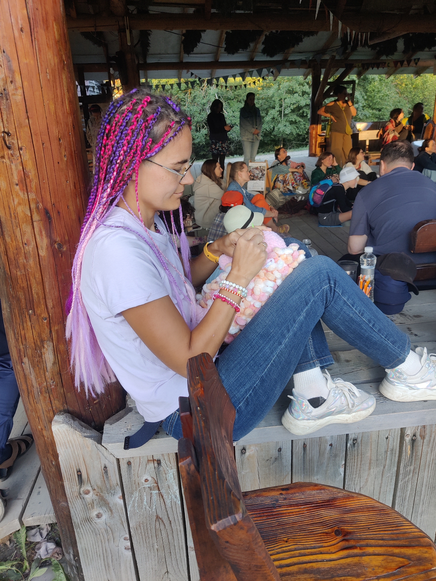 In Izky, Ukraine, a woman with pink and purple braided hair sits on a wooden bench, engaged in knitting a pink and white fluffy object. She wears a light purple t-shirt, blue jeans, white socks, and silver sneakers. Several bracelets adorn her left wrist. She is positioned in the foreground, leaning against a rough wooden pillar. In the background, a group of people are gathered under a covered structure with a thatched roof, with trees and foliage visible beyond. Some individuals are seated on the floor, while others stand. The setting appears to be an outdoor gathering or event.