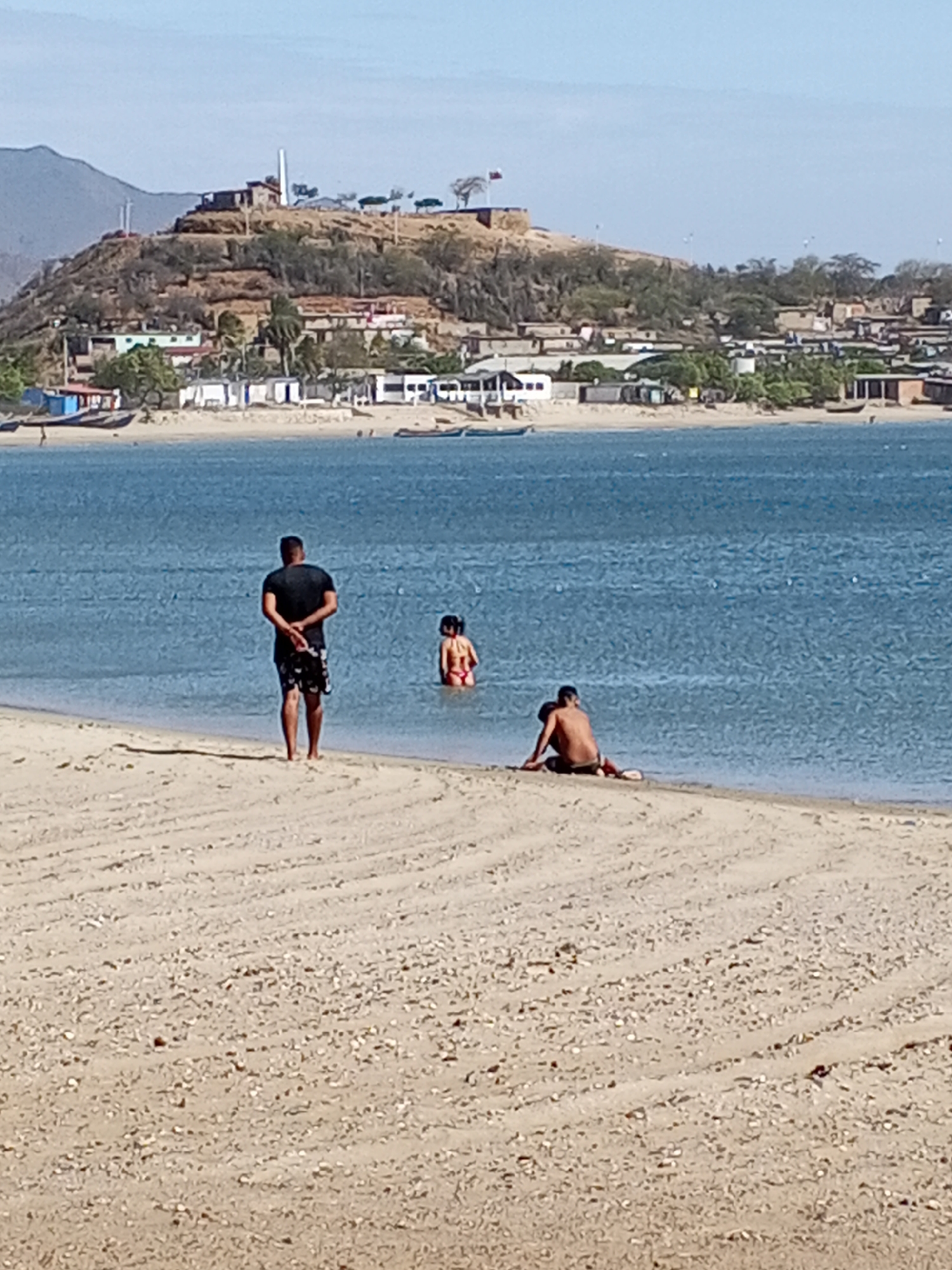 The image shows a beach scene in Juan Griego, Venezuela, with a view towards a hill topped by historical structures, including Fortin de la Galera. The sky is clear and light blue. The water is a calm, darker blue. In the foreground, there is a sandy beach.
On the beach and in the water: a man stands facing the water with his hands clasped behind his back, wearing a black shirt and patterned dark shorts; a child stands in the water, their back to the camera, wearing a red bathing suit; a person sits on the shoreline.
In the background: across the water is a hillside with buildings and vegetation, including a structure topped with a flagpole flying a national flag and the tower of Fortin de la Galera. The landscape includes low-lying houses and trees.