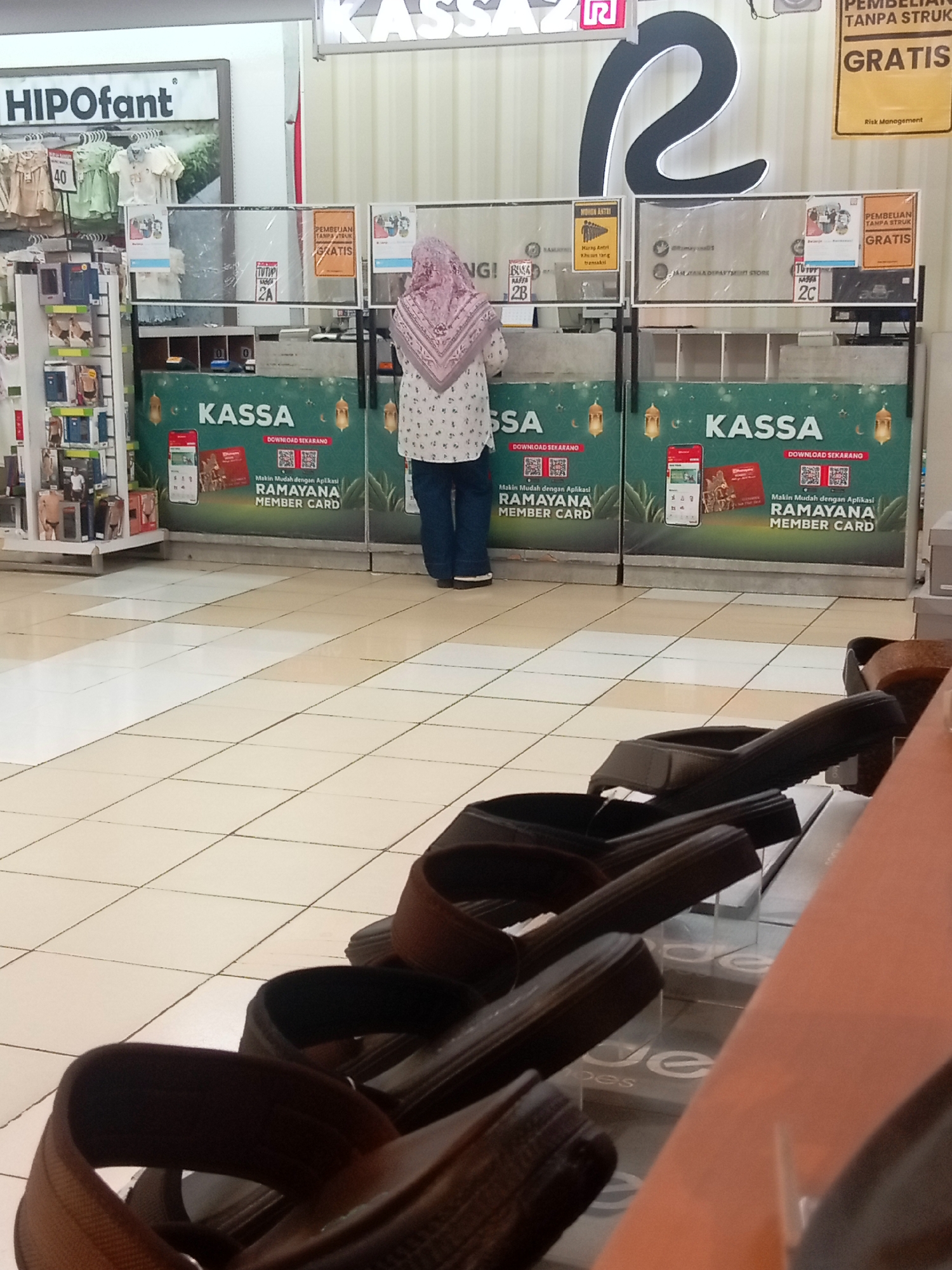 The image shows a scene inside a department store in Batam, Indonesia, likely at the Ramayana department store, given the prominent "KASSA" signs and "RAMAYANA MEMBER CARD" promotions. The foreground features a display of dark-colored sandals, some of which are casually placed on shelves. The middle ground shows a row of cashier counters, with two counters clearly visible. One counter has a "TUTUP" (closed) sign and a number "2A," while another has "2C." A woman wearing a pink hijab and a patterned white blouse, along with blue jeans, is standing at one of the counters, facing away from the camera. Behind the counters, there are promotional posters for "KASSA" and downloading the "RAMAYANA MEMBER CARD" application, featuring images of smartphones and membership cards. To the left of the counters, there's a display of children's clothing and what appears to be a retail display shelf with various items. The floor is tiled in a beige and white pattern. The overall environment suggests a retail setting during operating hours.