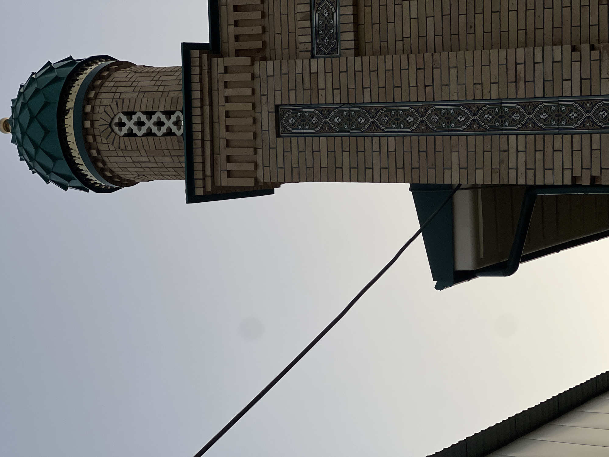The image shows the upper part of a mosque minaret and adjacent building in Tashkent, Uzbekistan. The minaret is constructed from light-colored bricks with decorative elements. It features a green, faceted dome topped with a golden finial. Below the dome, there is a band of intricately patterned tiles and a section with a geometric openwork design. The building wall also has decorative tilework. To the left of the minaret, a green-tiled roofline and a gutter are visible, along with the corner of another building with a corrugated metal facade. A black cable stretches diagonally across the sky, which is overcast and light gray. The photograph is taken from a low angle, looking upwards at the architecture against the sky. There are no people or animals present in the image. The scene suggests an architectural detail of a religious building in an urban setting.